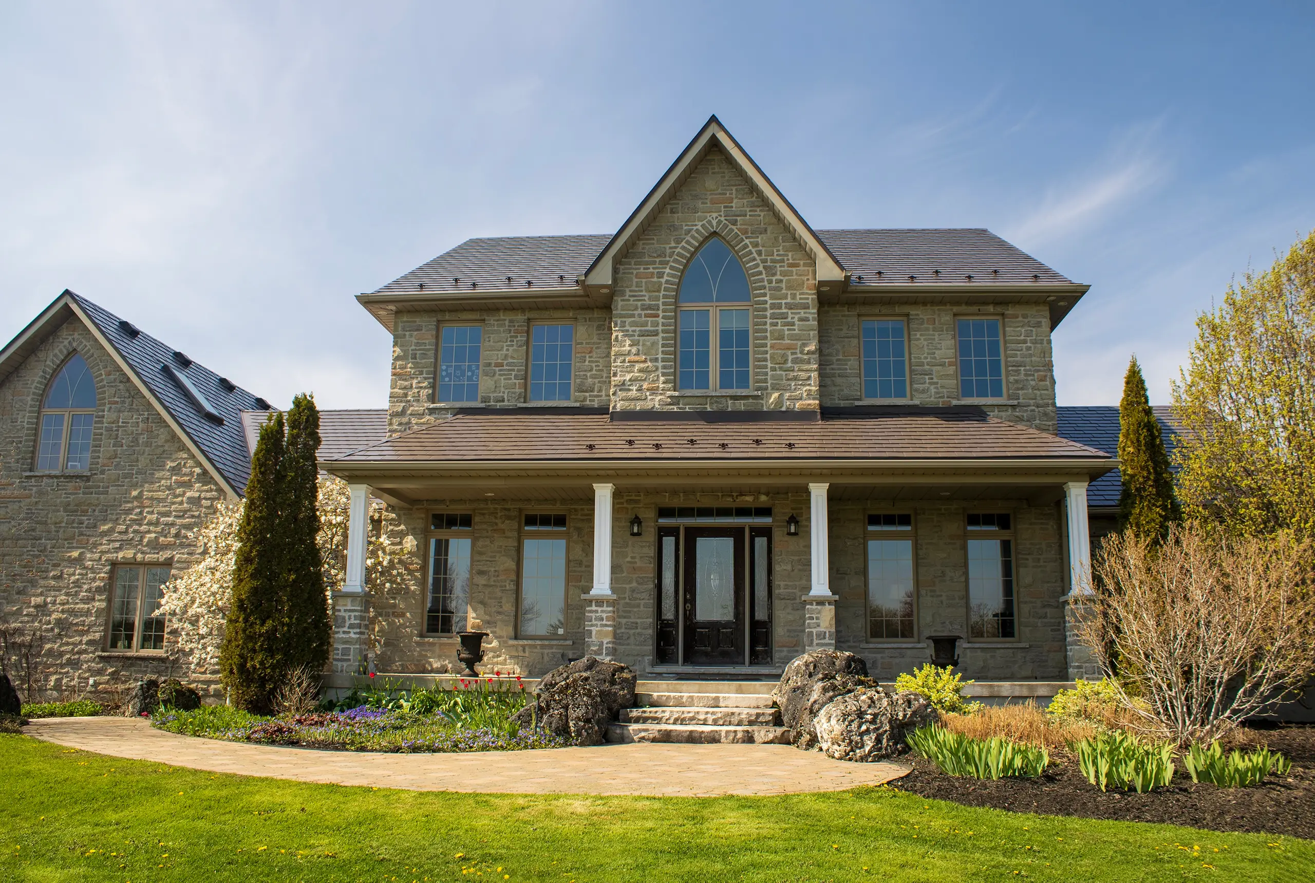 Two-storey Tudor-style stone home in Ariss, ON featuring Interlock Tudor Brown aluminum slate metal roofing with steep multi-pitch gables and Gothic arch window.