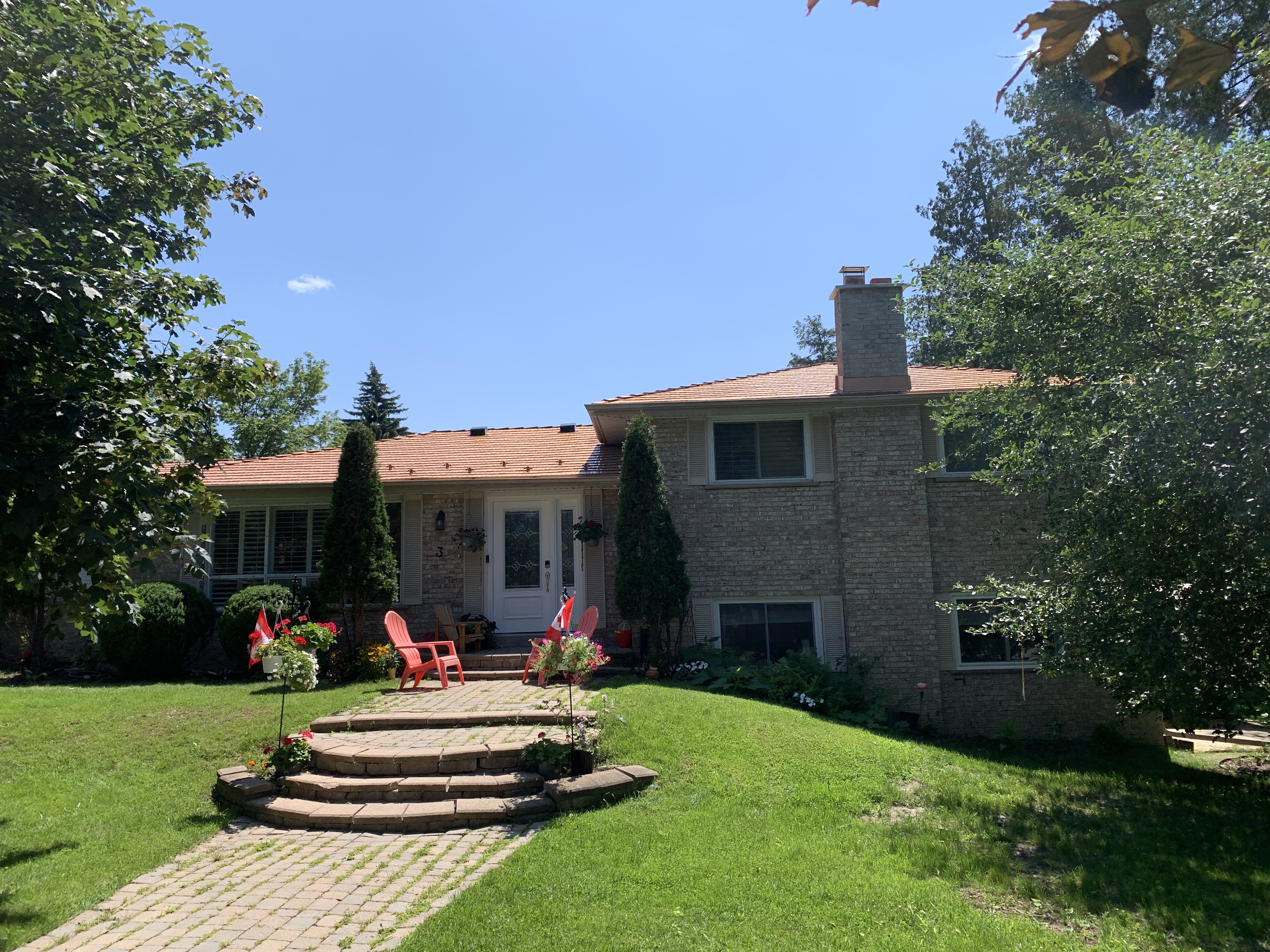 Front elevation of a 1.5-storey brick home in Aurora, ON featuring Interlock Copper Penny cedar shingle metal roofing with a chimney and mature tree surround.