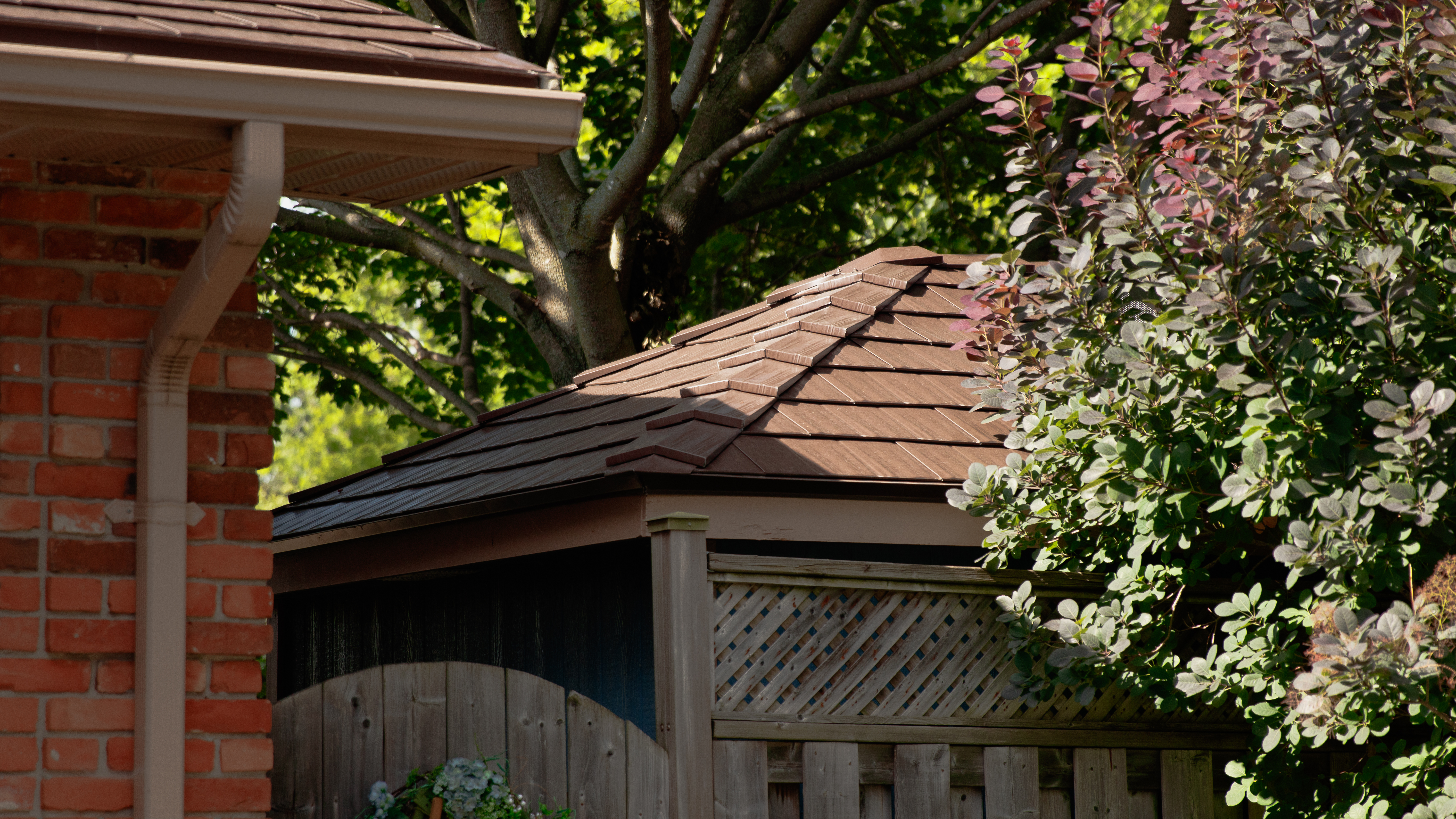 Interlock Colonial Brown cedar shingle aluminum hip roof on a small gazebo or hot tub enclosure beside a brick home, surrounded by lush garden greenery.