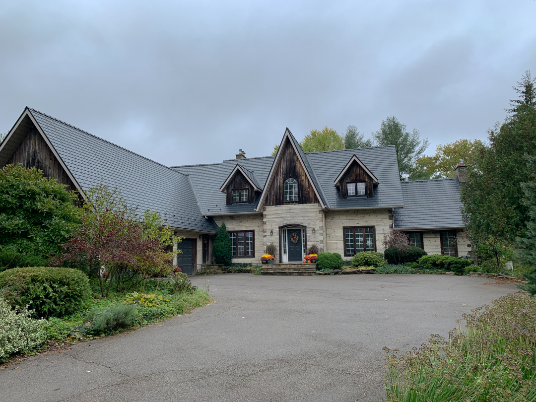 Front elevation of a Tudor-style stone home in St. George, ON featuring a steeply pitched Interlock aluminum slate roof in charcoal black with multiple dormers and complex valleys.