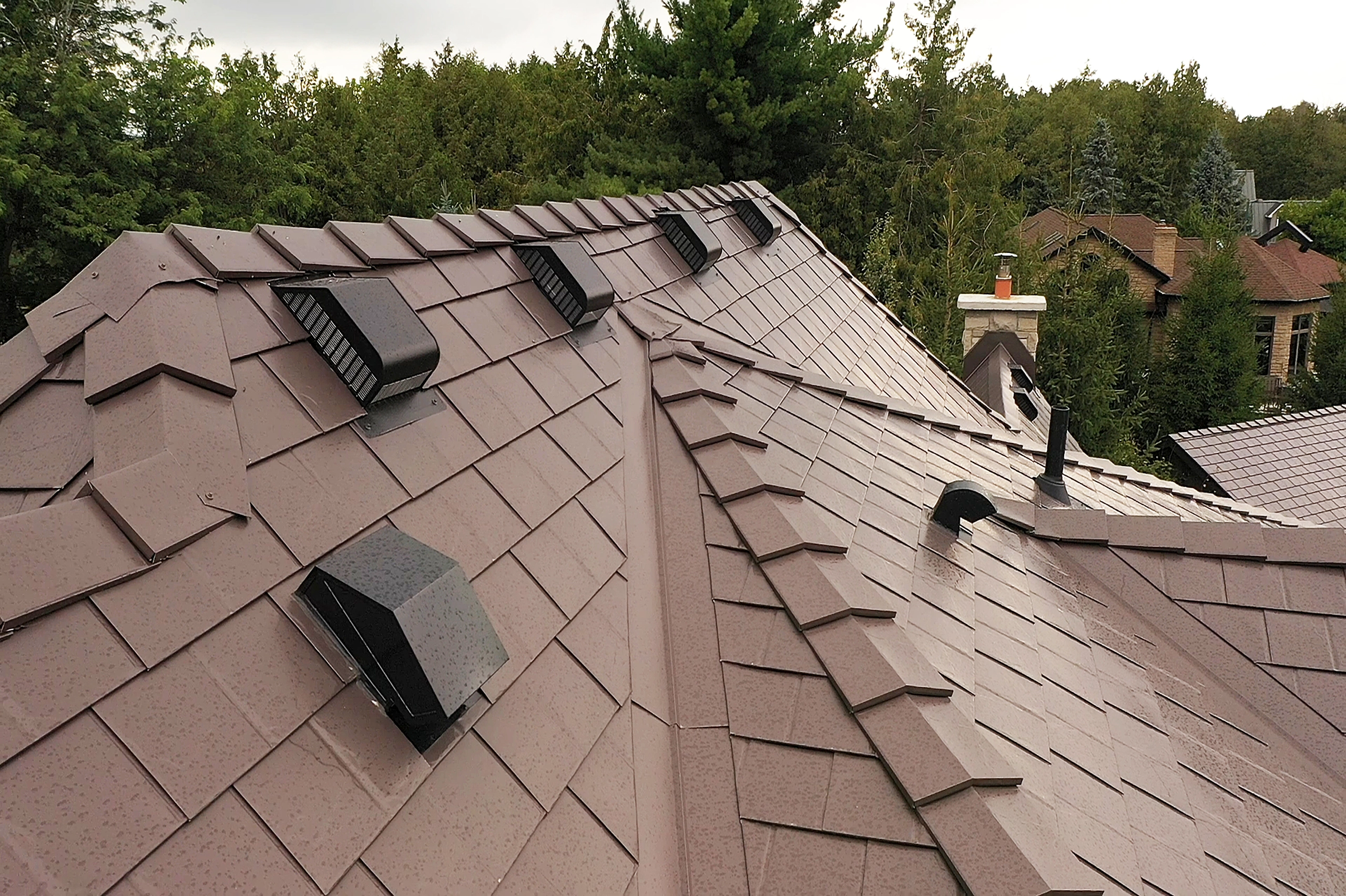 Complex Tudor Brown Interlock aluminum slate metal roof with multiple valleys, roof louvers, vent pipes, and ridge caps on a suburban forested home.