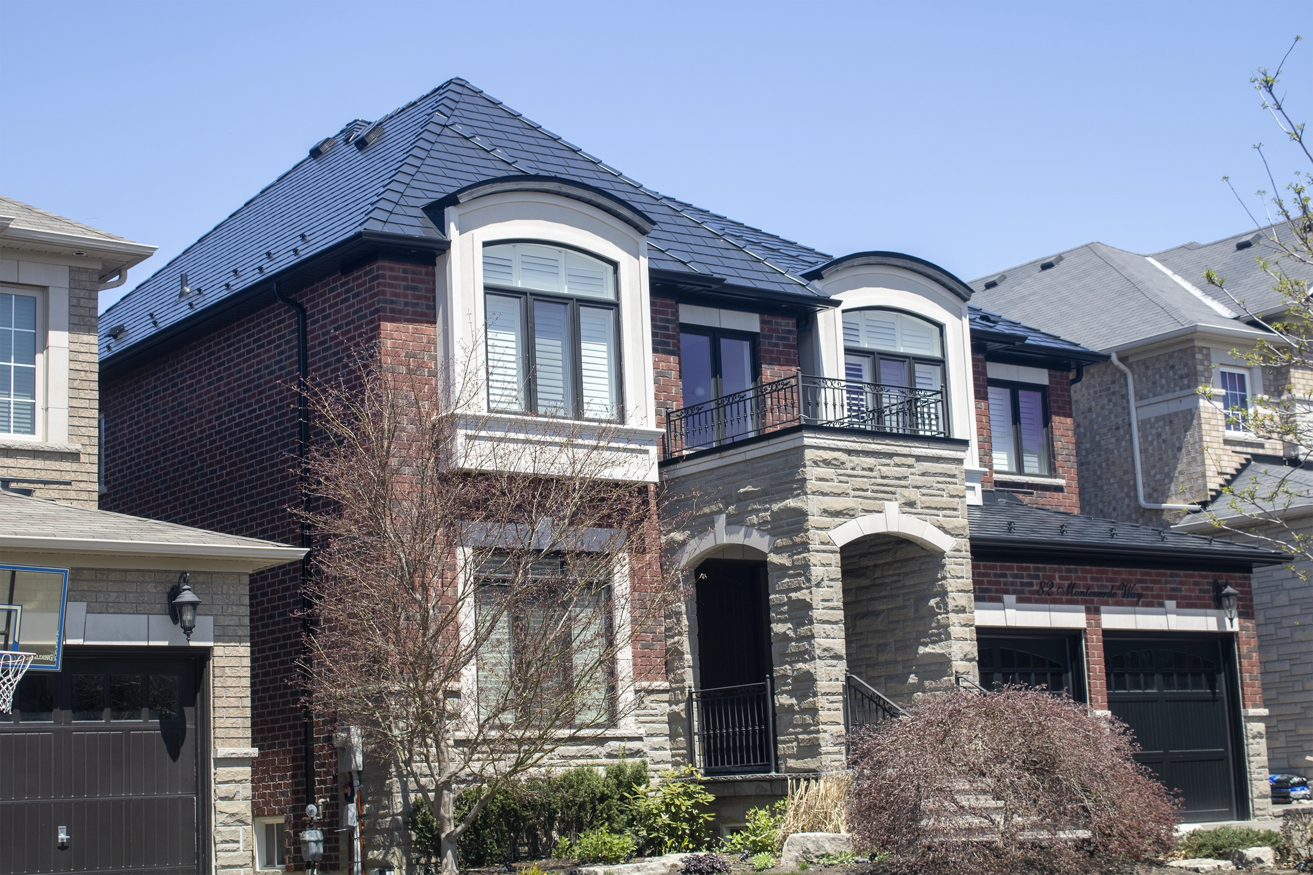 Two-storey brick and stone Colonial home in Woodbridge, ON with a complex multi-hip black Interlock aluminum slate metal roof and wrought-iron balcony.