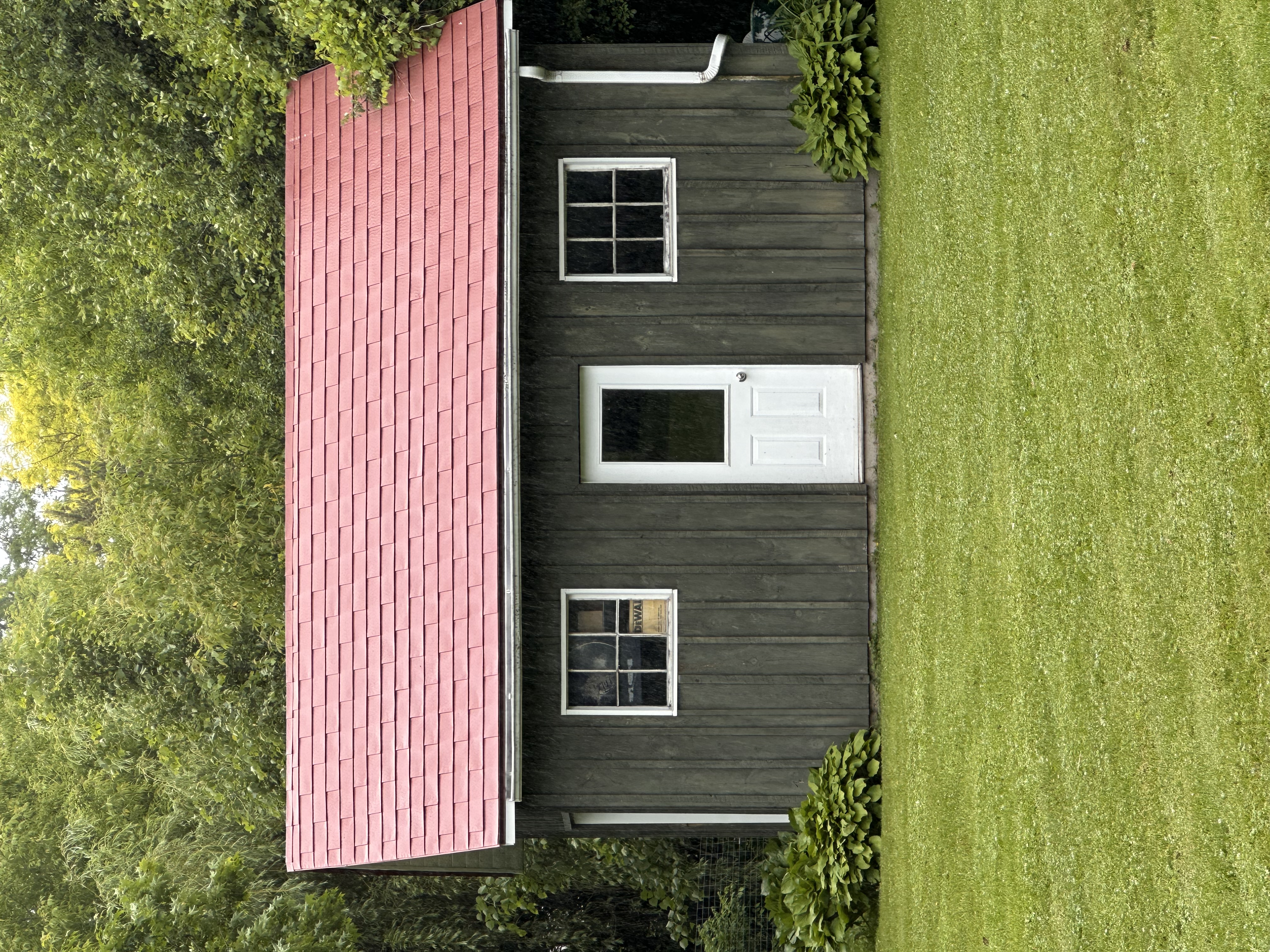 Front elevation of a rustic wood-clad outbuilding with a Tile Red Interlock aluminum shingle gable roof, white door, and twin windows in Almonte, ON.