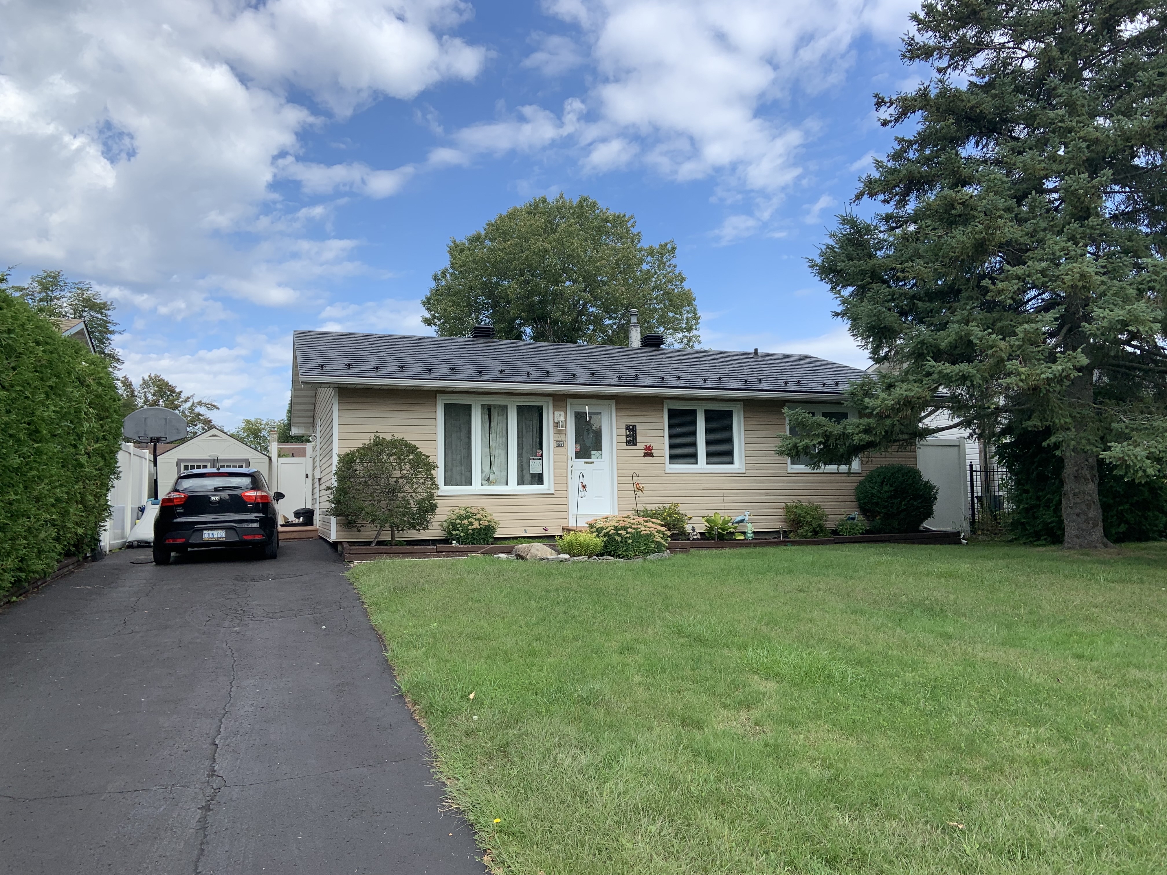 Front elevation of a single-storey ranch home in Ottawa with a black Interlock aluminum slate metal roof, beige siding, and mature trees.