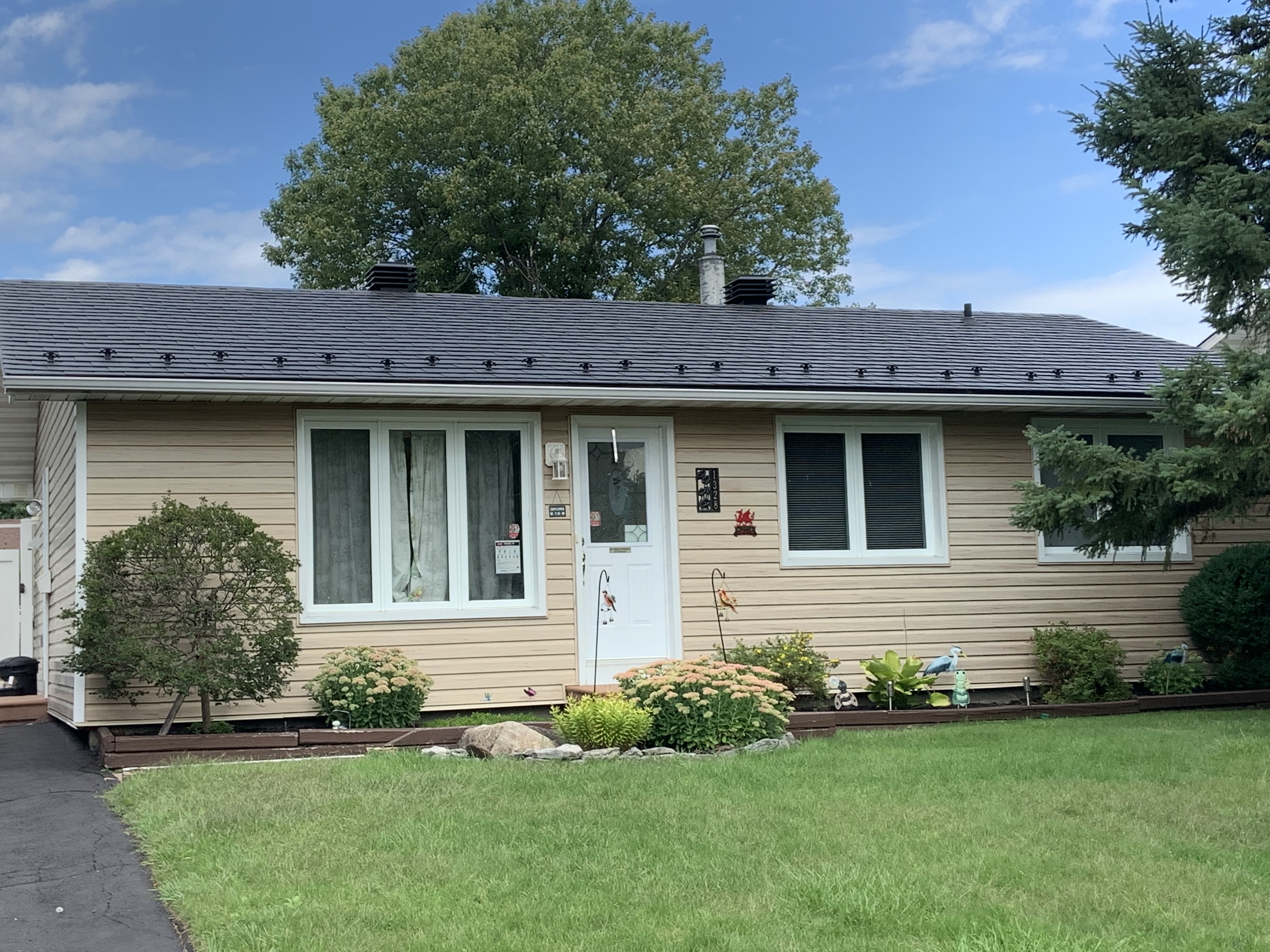 Single-story ranch home in Ottawa with Interlock black aluminum slate metal roof, low-pitch simple gable, tan vinyl siding, and manicured front garden.