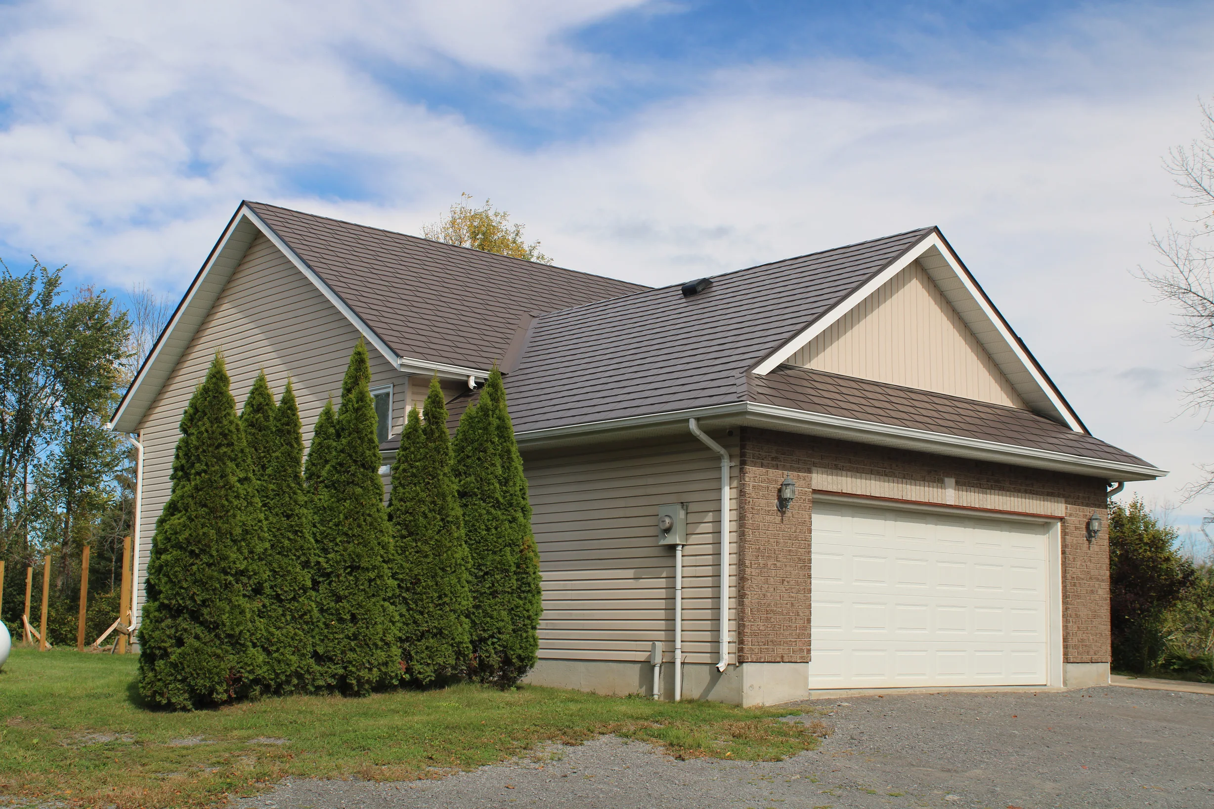 Tudor Brown Interlock Slate aluminum roof on a tan house with a brown metal roof among tall trees, Kingston, Ontario