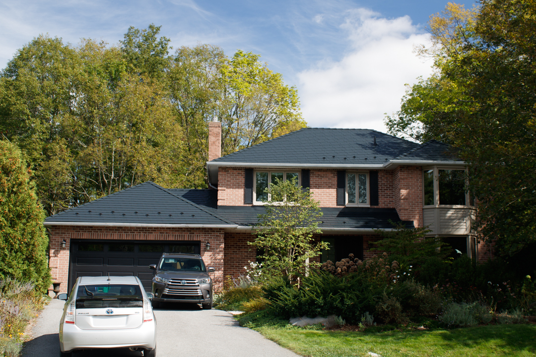 Two-storey brick Colonial home in Kingston, ON with a black Interlock aluminum slate metal roof, chimney, and lush tree-lined suburban setting.