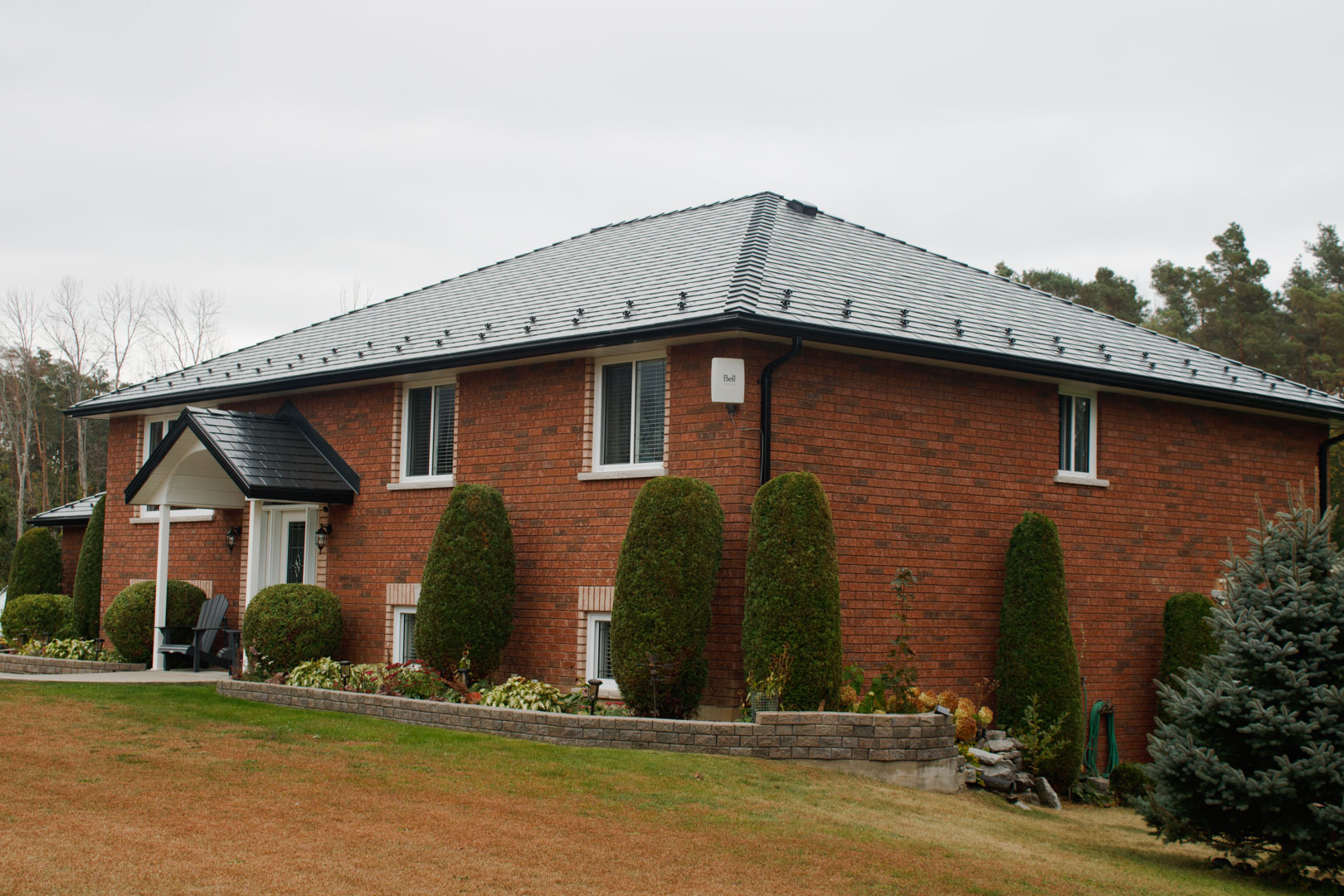 Red brick single-family home with a dark charcoal Interlock aluminum slate hip roof, front portico, and manicured evergreen landscaping in rural Ontario.