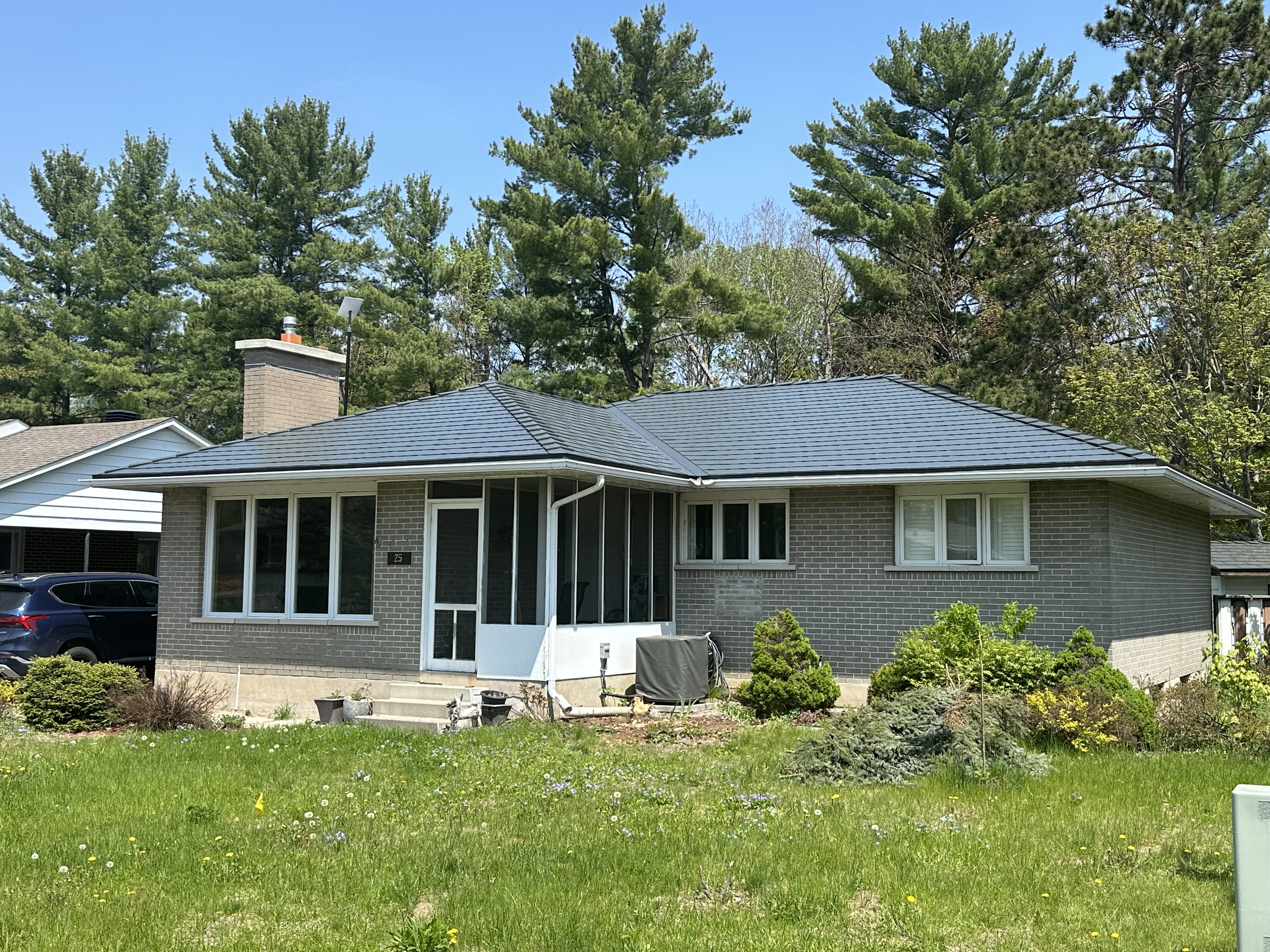 Single-storey ranch-style brick home with a deep charcoal Interlock aluminum slate hip roof, chimney, and screened front entry, set against tall pines.