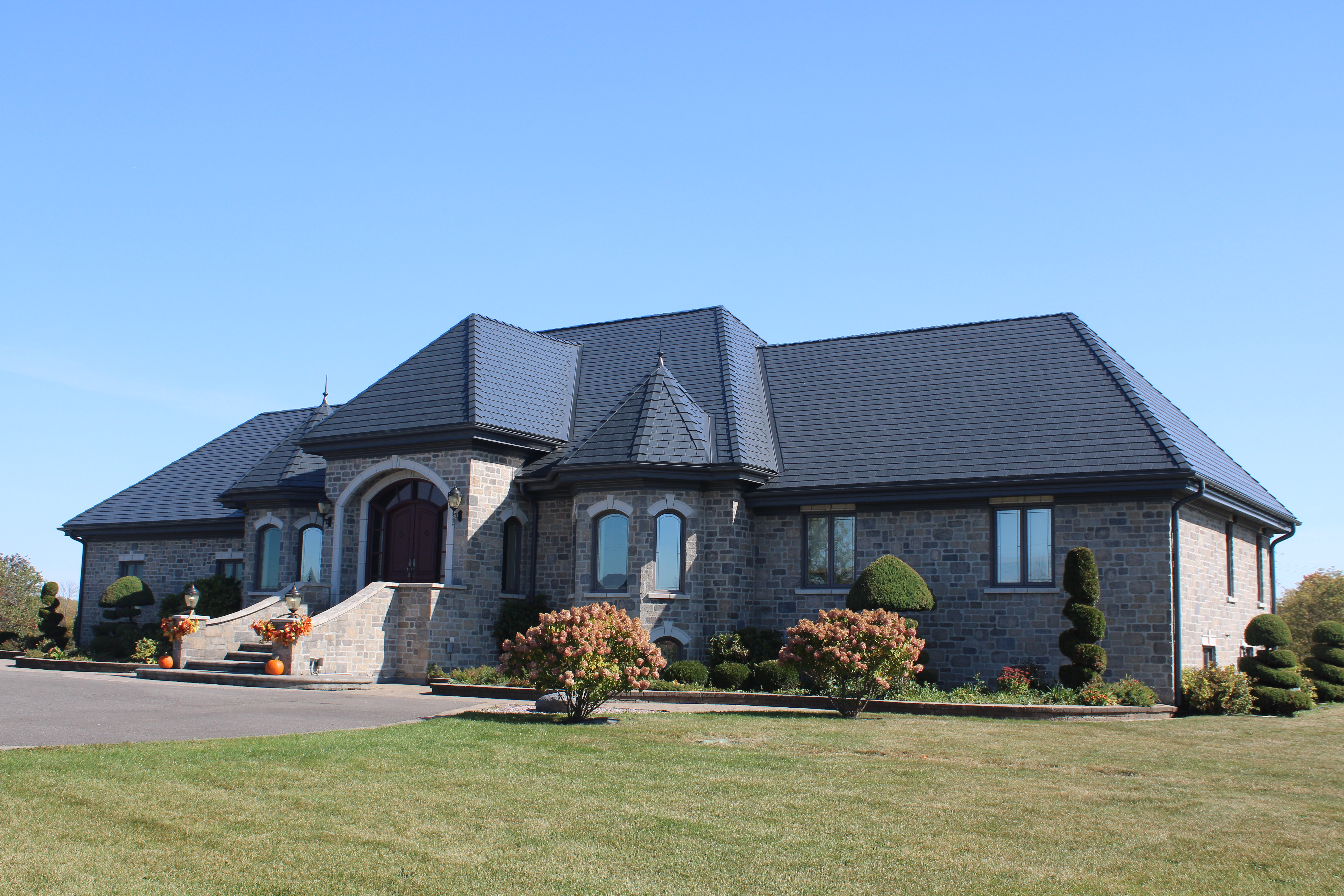 Large stone estate home in Alexandria ON with deep charcoal Interlock aluminum slate roof featuring a conical turret, multiple hip planes, and manicured topiary landscaping.