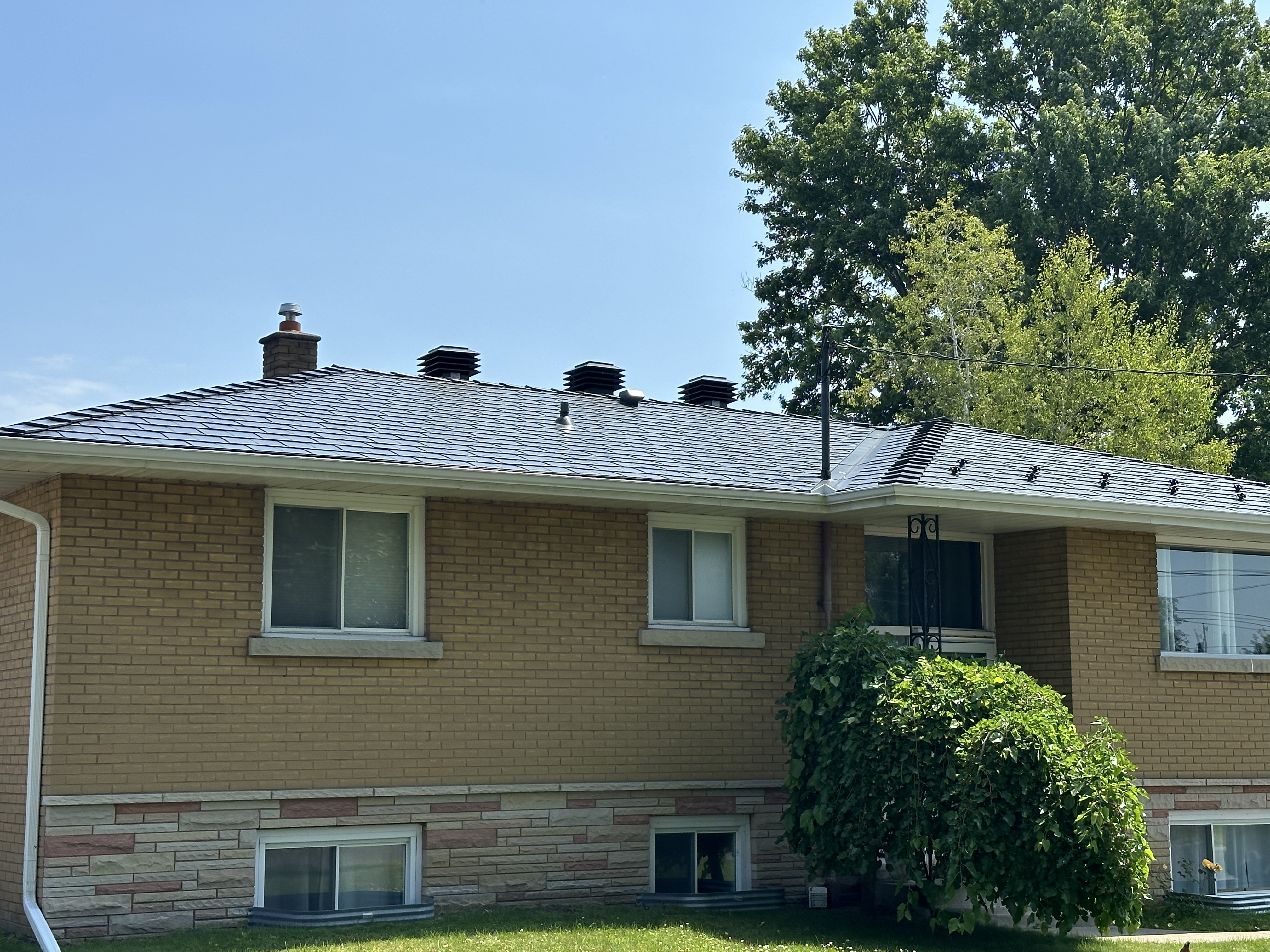Black Interlock aluminum slate metal roof on a single-storey ranch-style brick home in Cornwall, ON, featuring multiple roof louvers and a brick chimney.