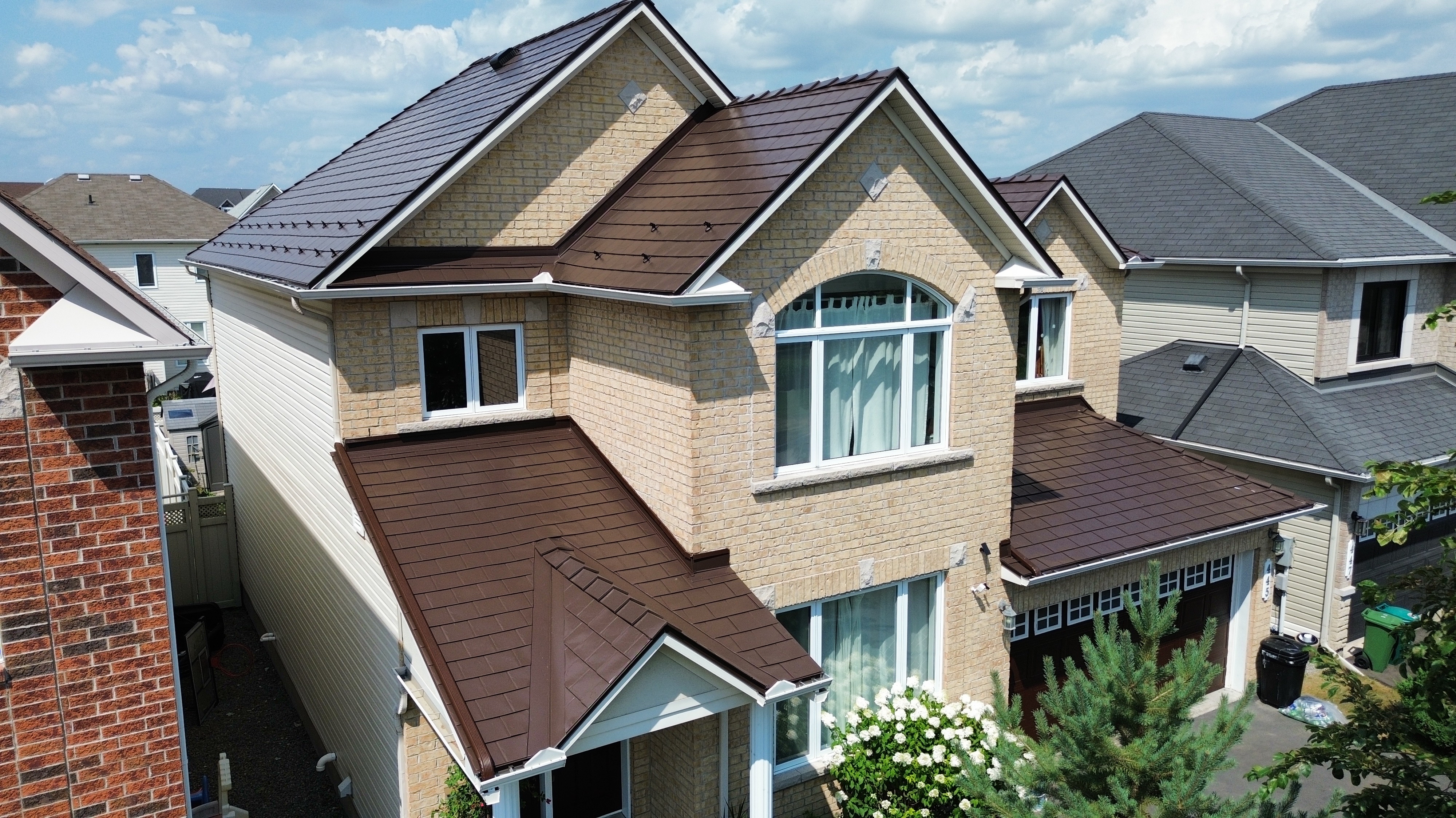 Aerial front view of a 2-storey suburban home with Interlock Colonial Brown aluminum slate metal roofing, complex multi-gable roof, and light brick exterior.