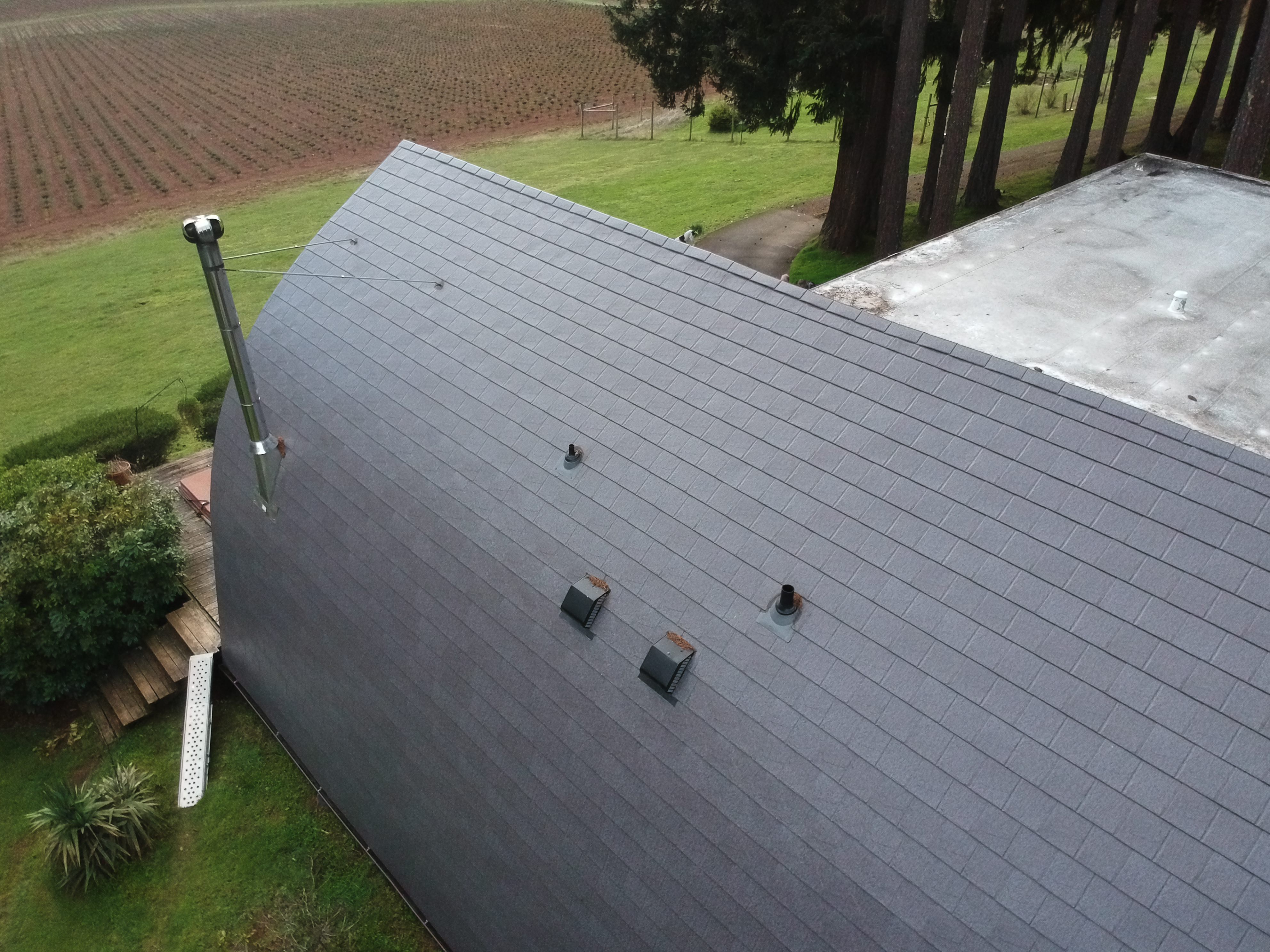 Aerial view of a curved Interlock aluminum slate roof in weathered gray on a rural Oregon farmhouse, with vent pipes and roof louvers visible.