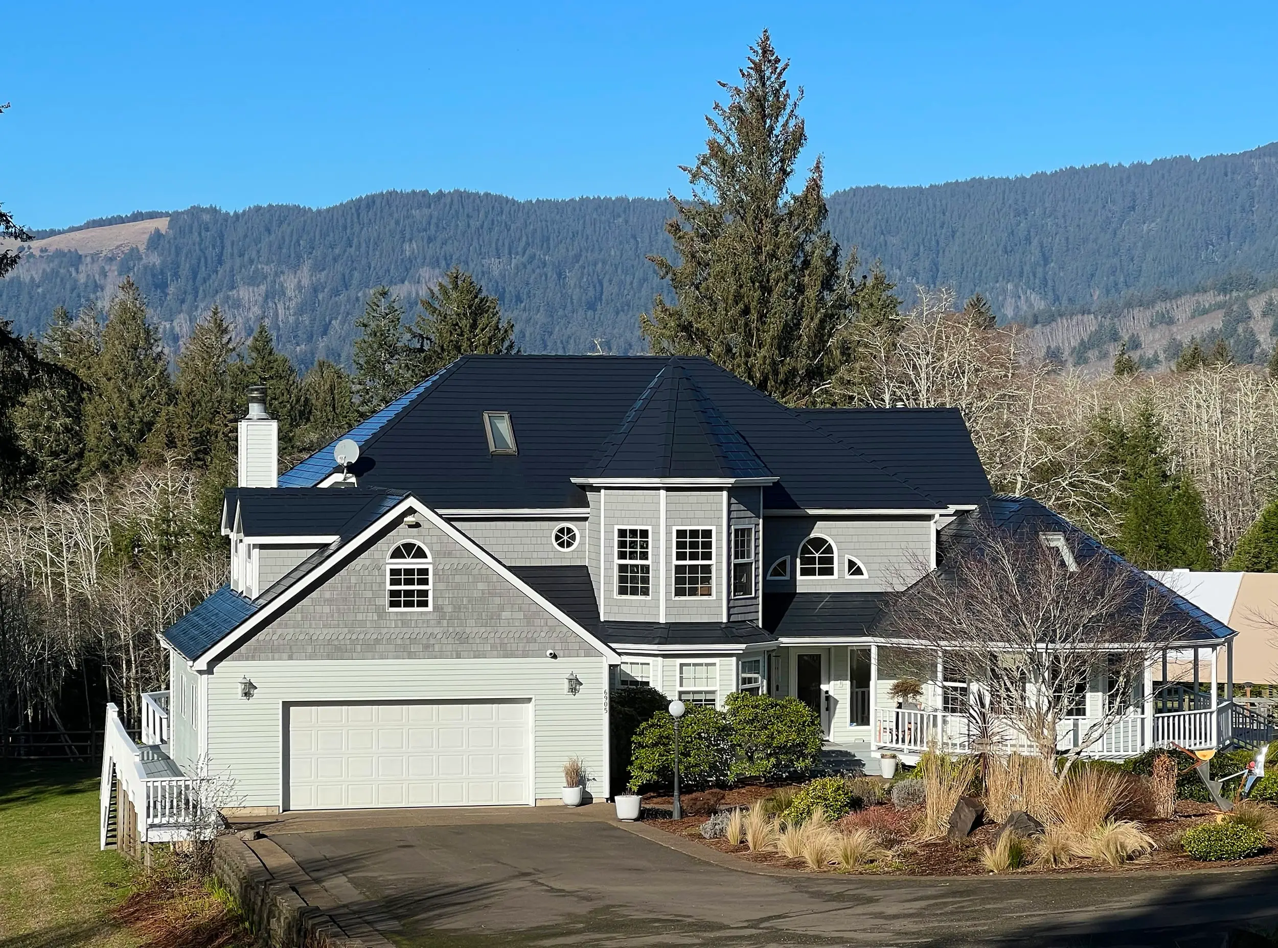 Two-story gray shingle-sided home in Otis, OR with a complex black Interlock aluminum slate metal roof, chimney, skylight, and mountain backdrop.