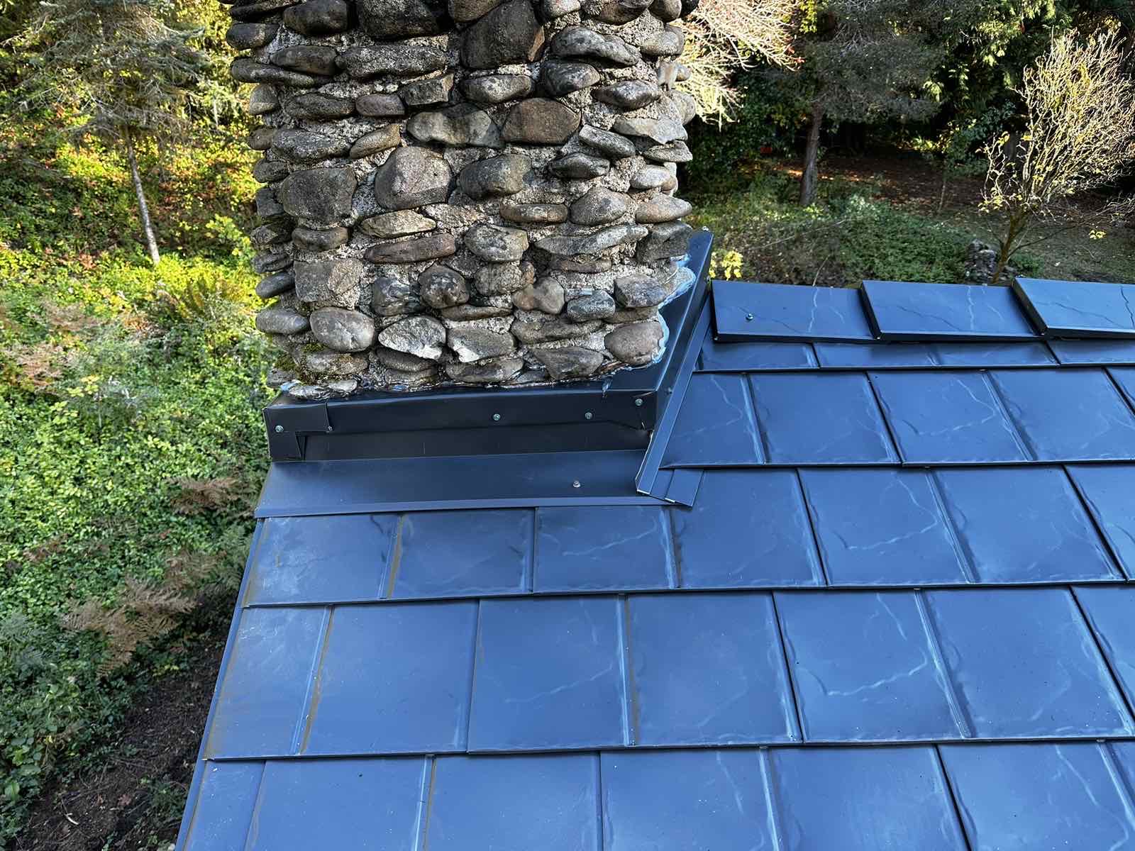 Close-up of black Interlock aluminum slate roof panels with custom metal step flashing around a rustic round-stone chimney, surrounded by forested landscape.