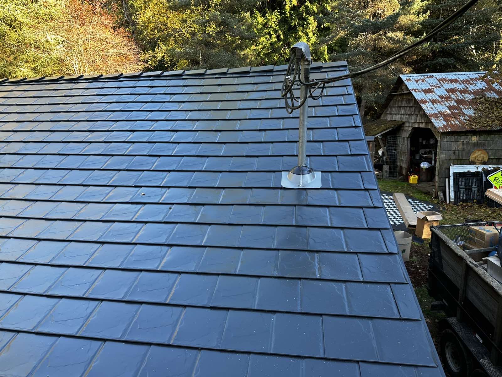 Black Interlock aluminum slate metal roof on a rural cottage in Alsea, OR, showing uniform tile layout, vent pipe flashing, and forested backdrop.
