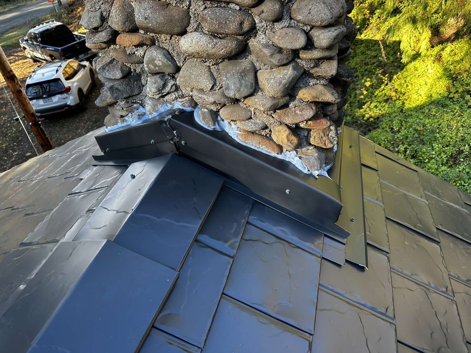 Close-up of black Interlock aluminum slate roof panels with custom metal step flashing around a river-rock stone chimney, Alsea OR.