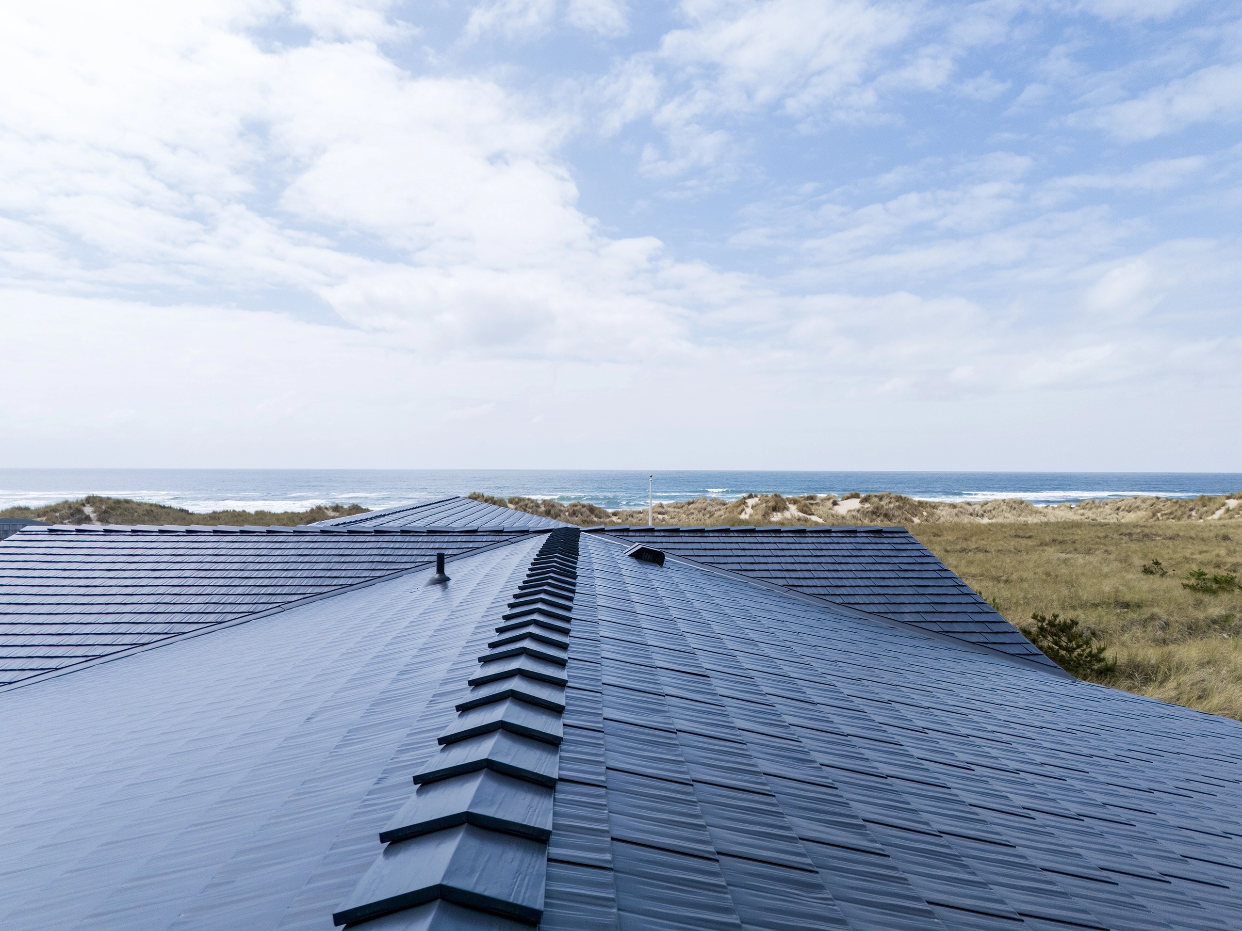 Aerial view of a black Interlock aluminum cedar-shingle metal roof with ridge cap, valley, and vent pipe, overlooking Oregon coastal dunes and ocean.