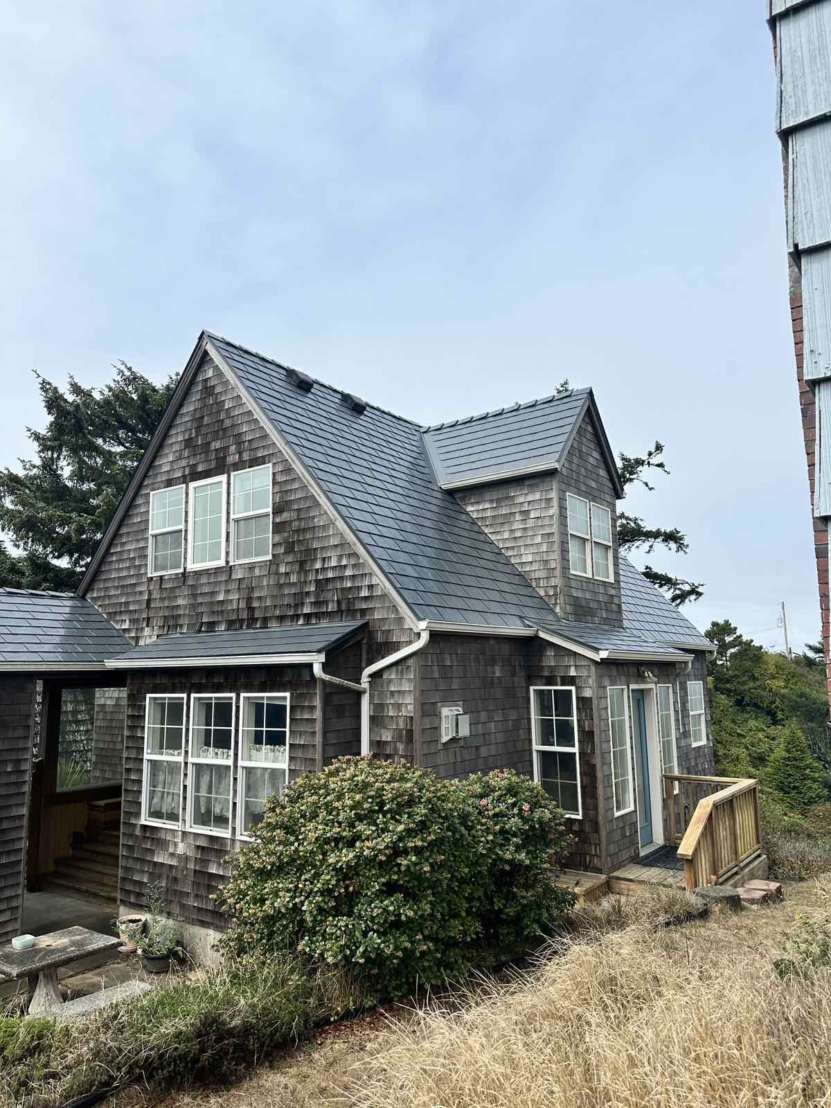 Two-story cedar-shingle coastal cottage in Newport, OR with a deep charcoal Interlock aluminum slate metal roof featuring multiple dormers and complex valleys.