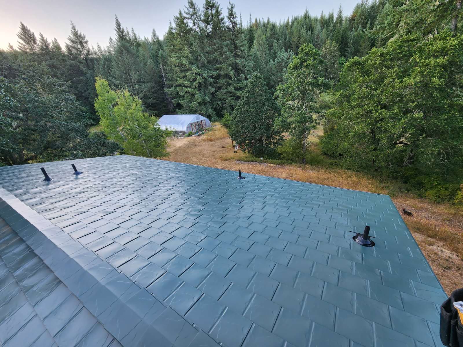 Aerial view of a hunter green Interlock aluminum slate metal roof with three vent pipes, surrounded by dense Pacific Northwest forest in Eugene, OR.