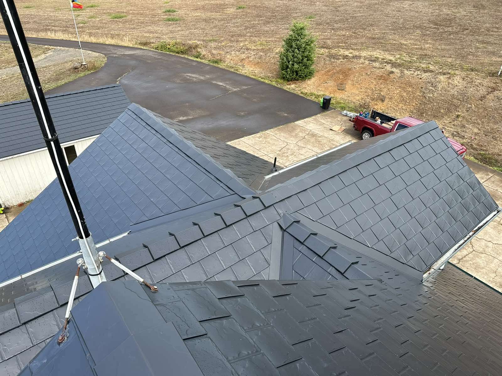 Aerial drone view of a complex multi-hip black Interlock aluminum slate metal roof with multiple valleys, snow guards, and ridge caps on a rural single-family home.