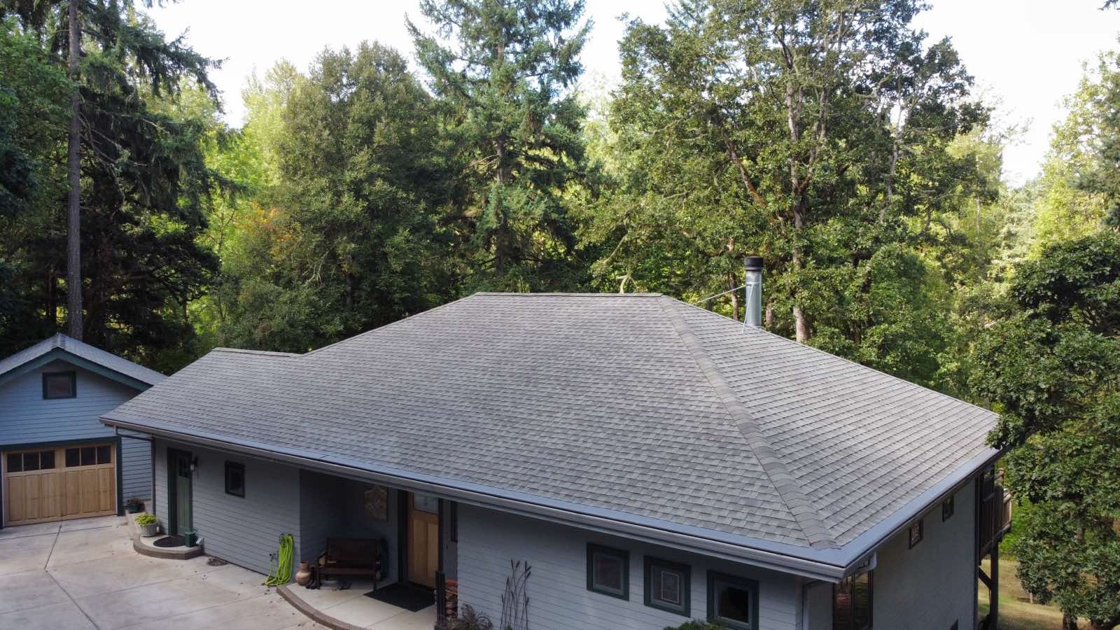 Aerial front view of a ranch-style home with Interlock Musket Grey aluminum slate metal roofing, hip roof, and wooded backdrop in Eugene, OR.