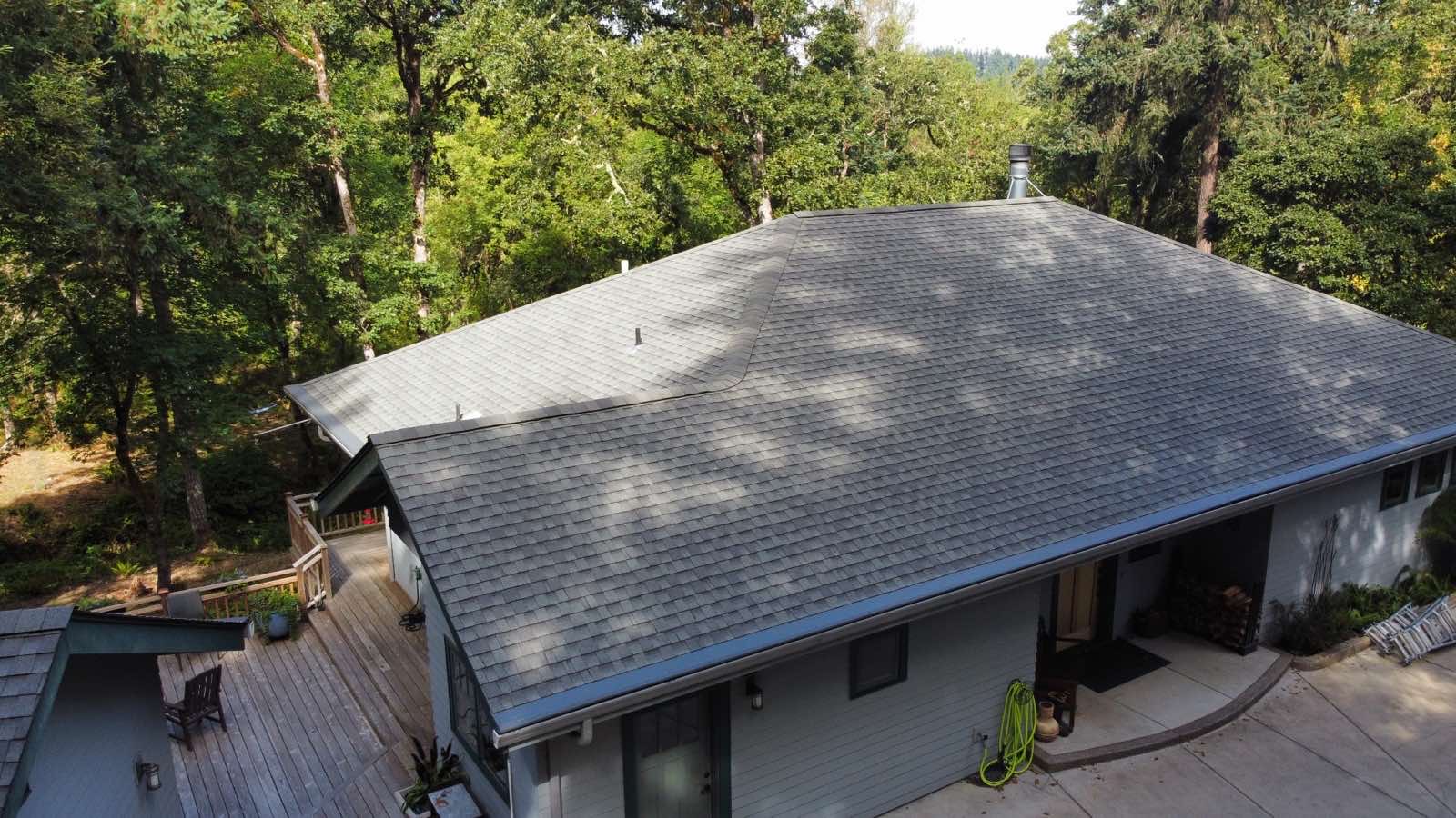 Aerial view of a ranch-style home in Eugene OR with an Interlock Musket Grey aluminum slate metal roof featuring hip and valley details surrounded by dense forest.