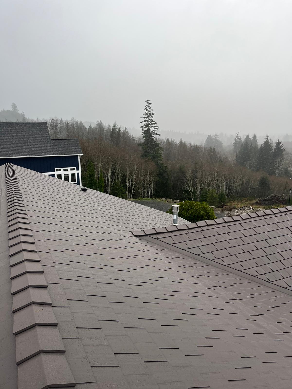 Tudor Brown Interlock aluminum slate metal roof with complex multi-hip planes, valley intersections, and vent pipe, set against a misty forested Oregon coast backdrop.