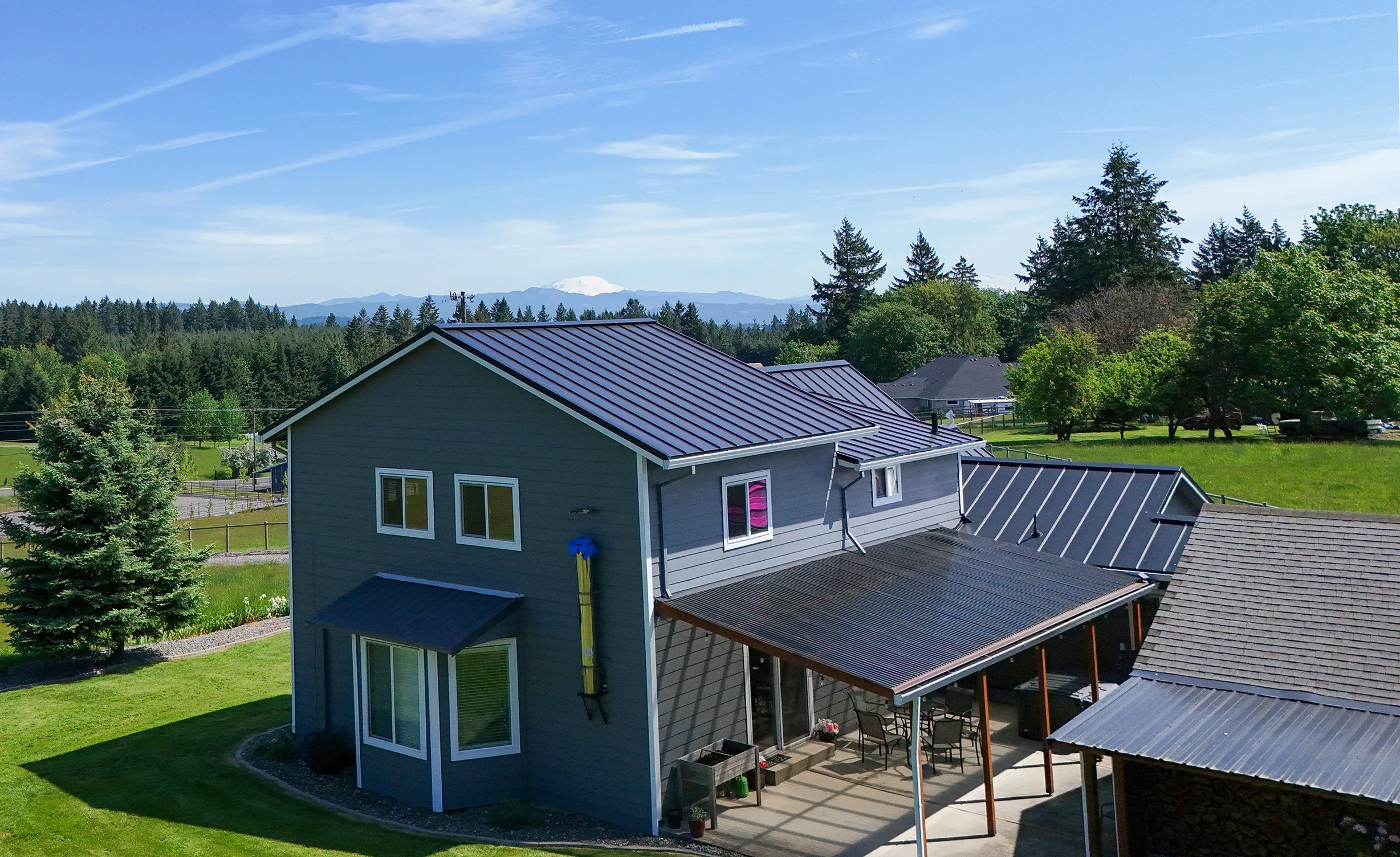 Black Interlock Standing Seam aluminum roof on a grey two-storey home with dormers among tall pines, Warren, Oregon
