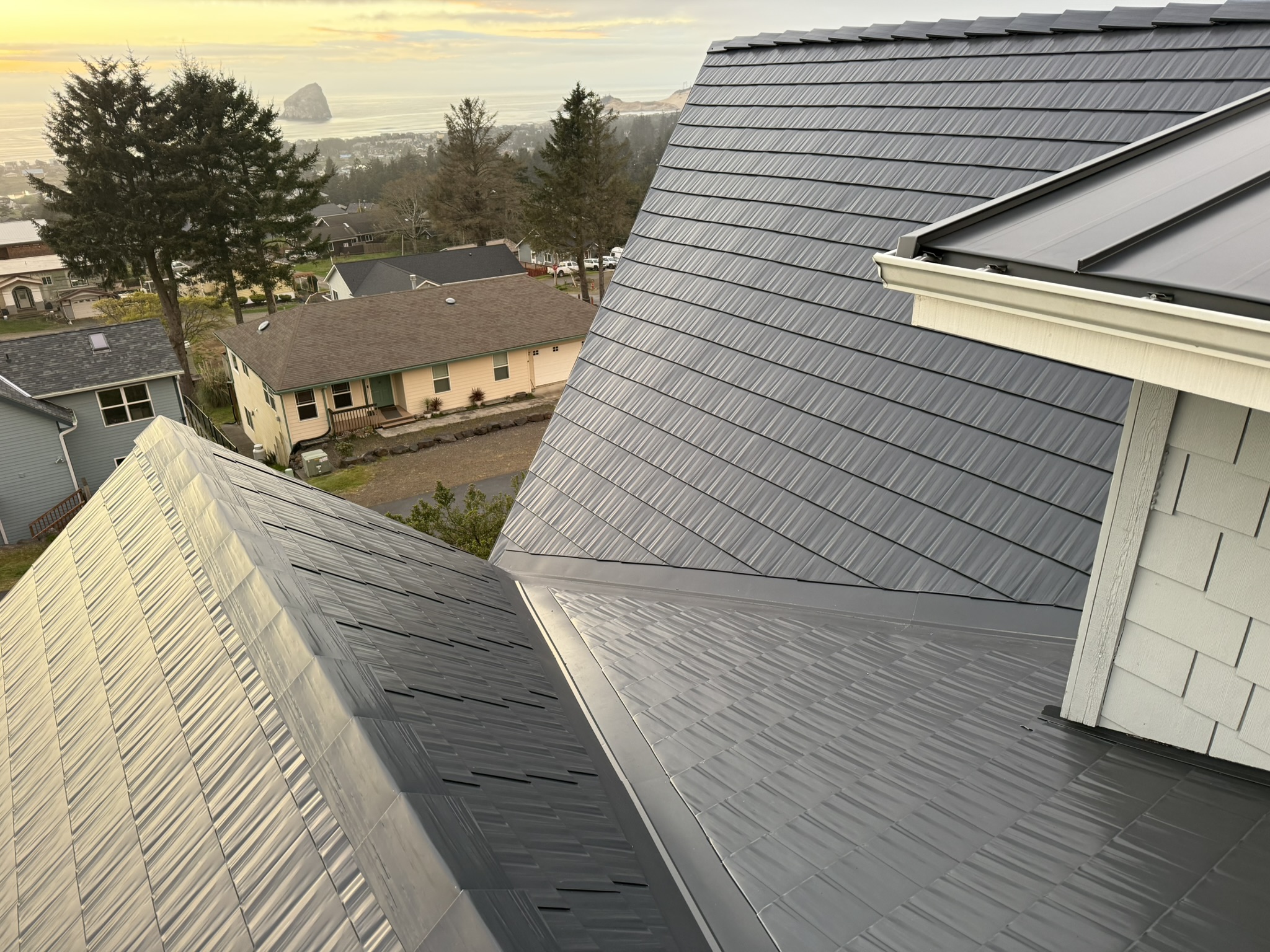 Aerial view of a complex multi-plane Interlock deep charcoal cedar shingle metal roof with valleys, overlooking a coastal Oregon town at golden hour.