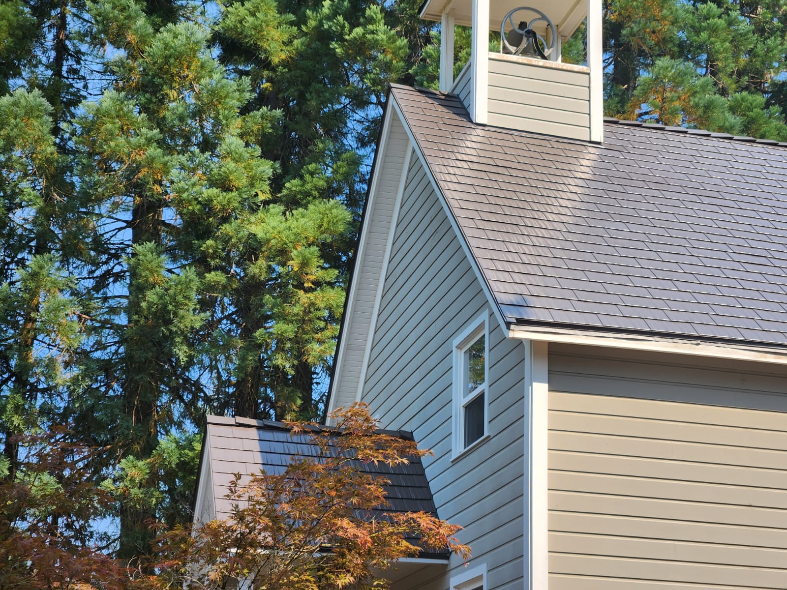 Steep-pitched Tudor Brown Interlock cedar shingle metal roof on a gray-sided church with bell tower cupola, set among tall Pacific Northwest conifers.