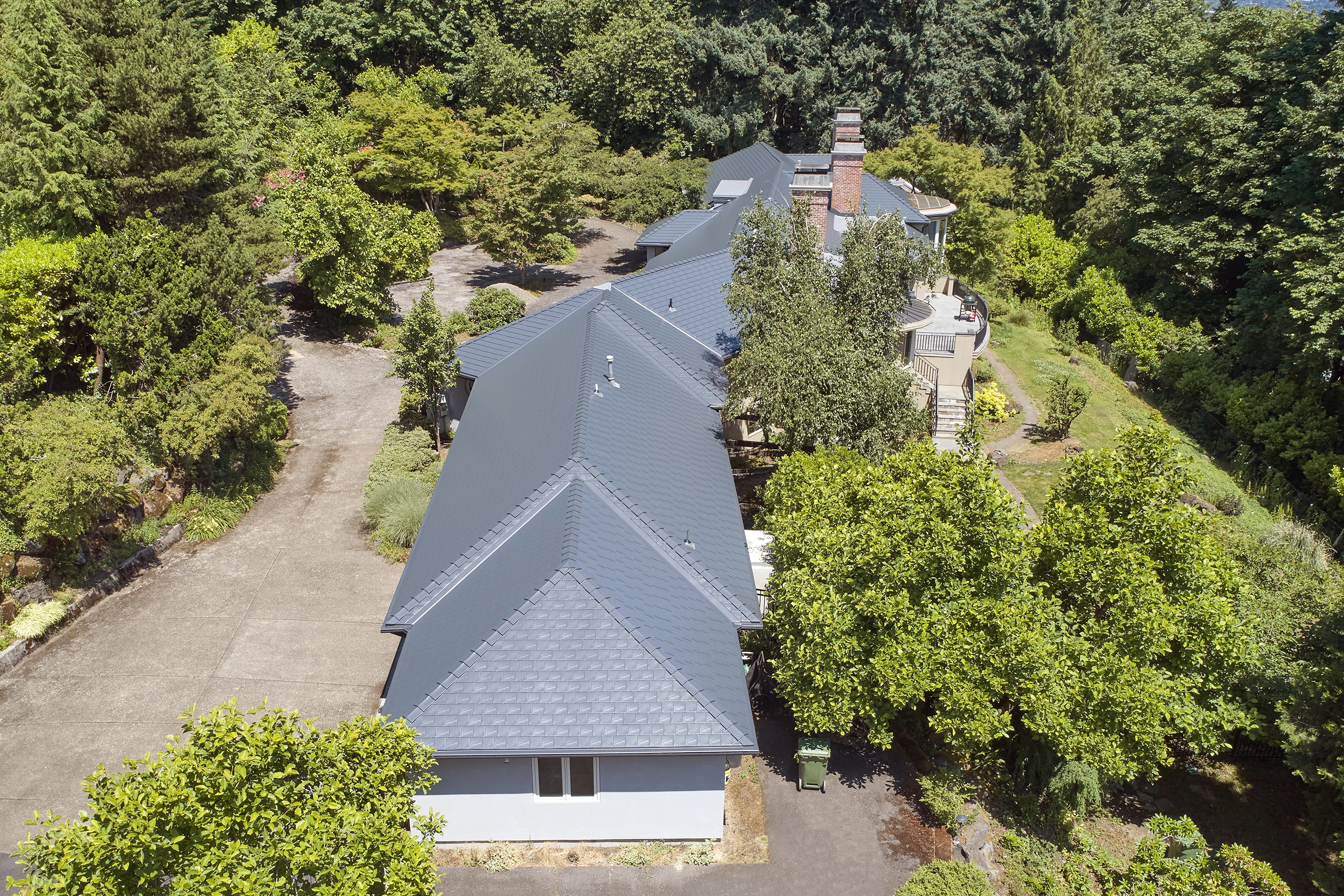 Aerial drone view of a sprawling single-family home with deep charcoal Interlock aluminum slate metal roofing featuring complex multi-hip geometry, valleys, and a brick chimney amid dense Portland foliage.