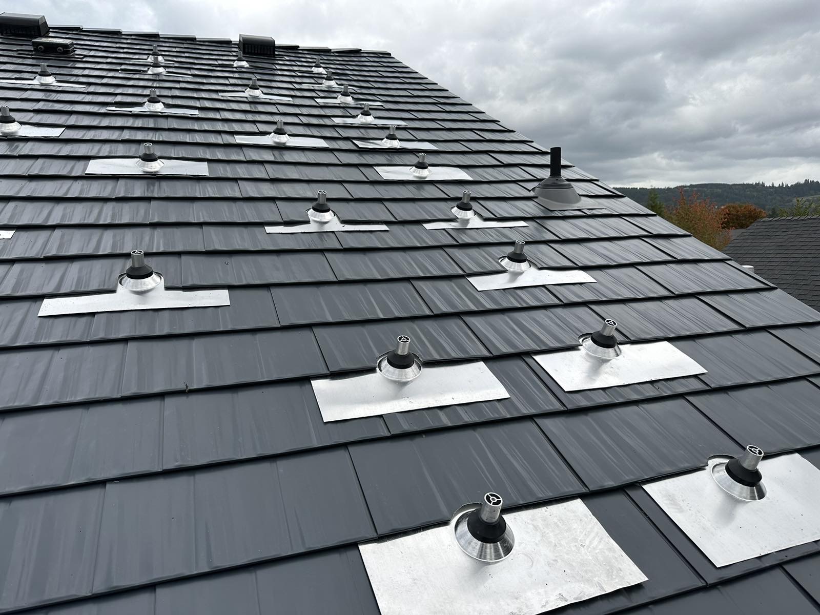 Close-up of deep charcoal Interlock aluminum cedar shingle metal roof with multiple aluminum-flashed vent pipe penetrations on a hip roof.