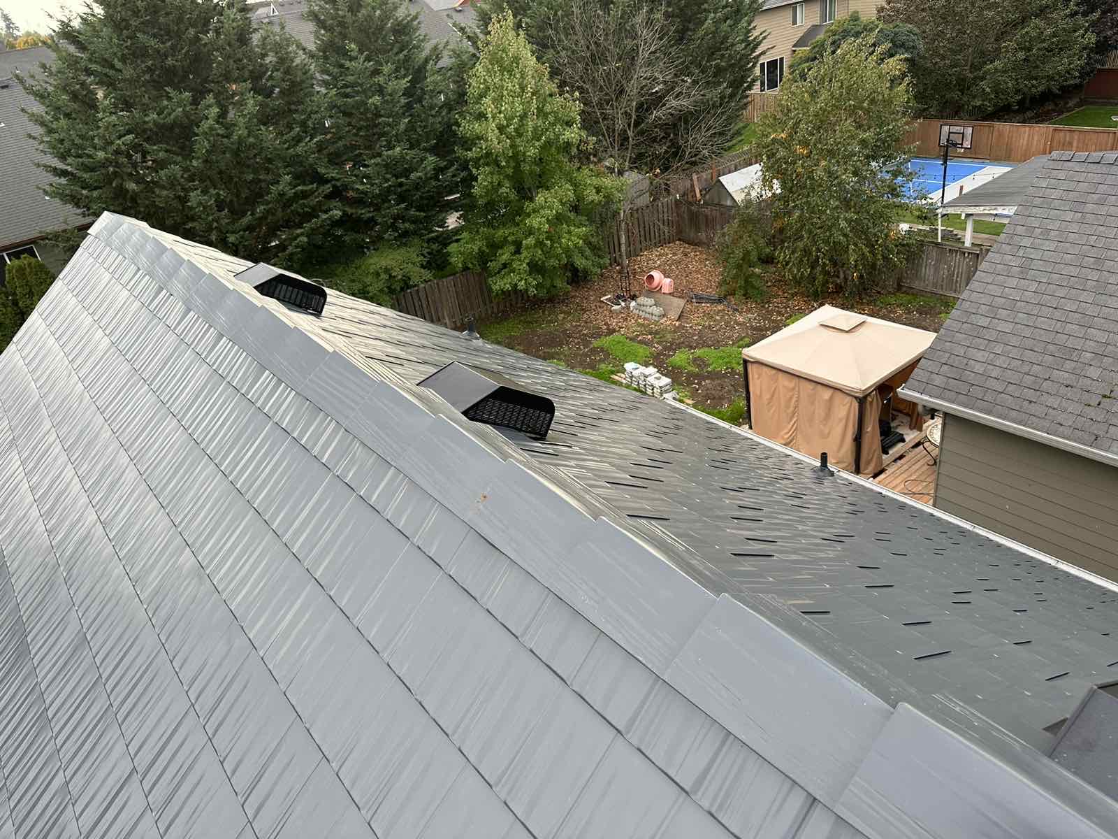 Aerial ridge-line view of a deep charcoal Interlock aluminum cedar shingle metal roof with ridge cap and roof louvers on a suburban two-story home.