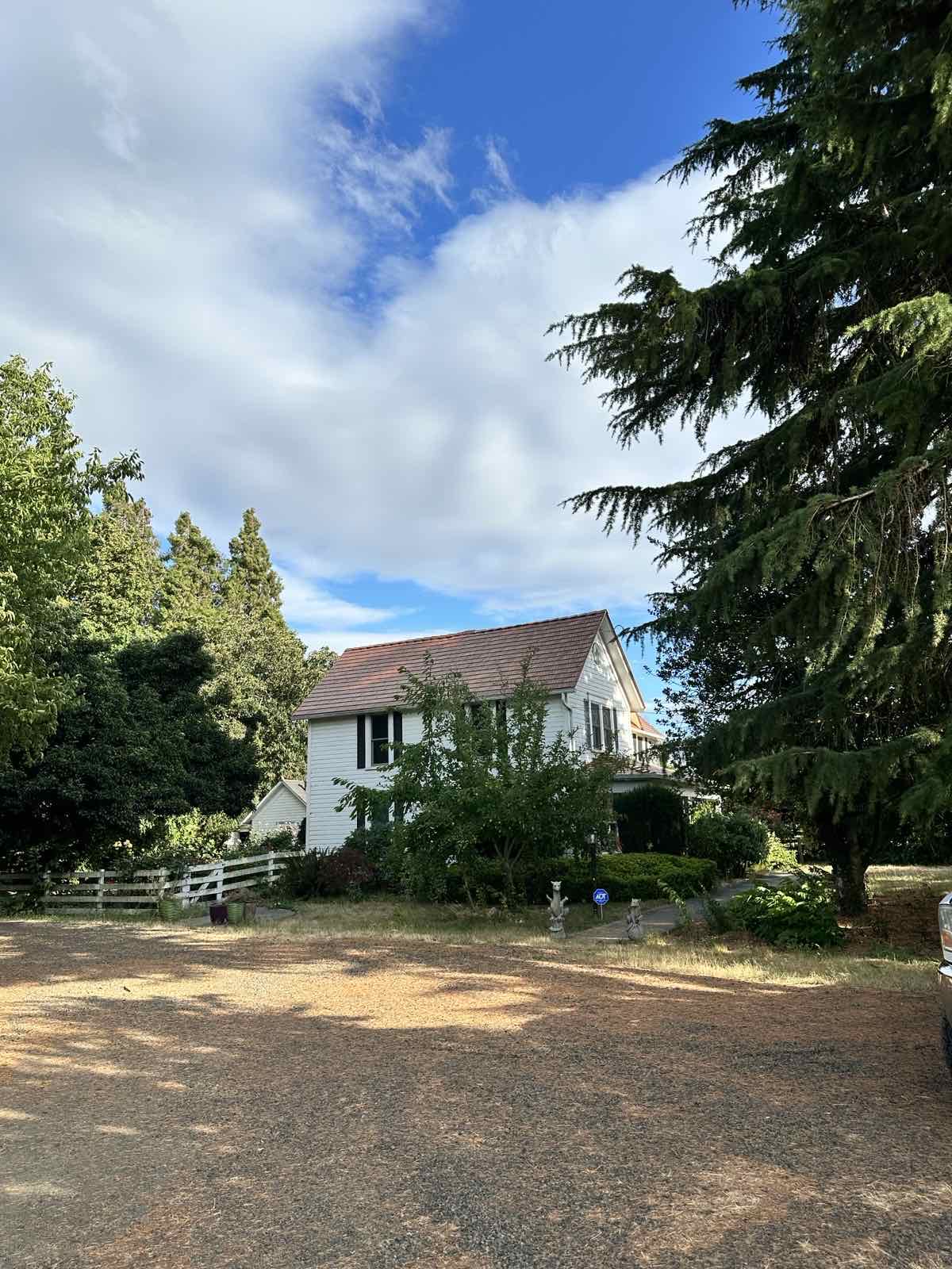 White two-story farmhouse with Interlock Copper Penny cedar shingle metal roof, surrounded by mature evergreen and deciduous trees in rural McMinnville, OR.