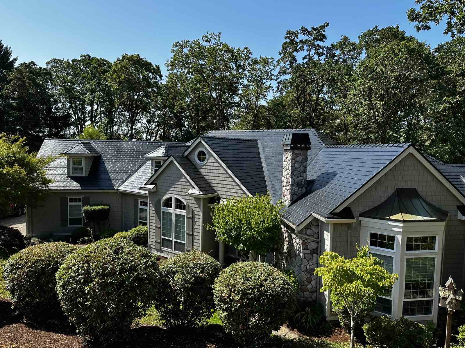 Aerial front view of a Craftsman home in Corvallis, OR with a complex black Interlock aluminum slate metal roof, stone chimney, and dormers.