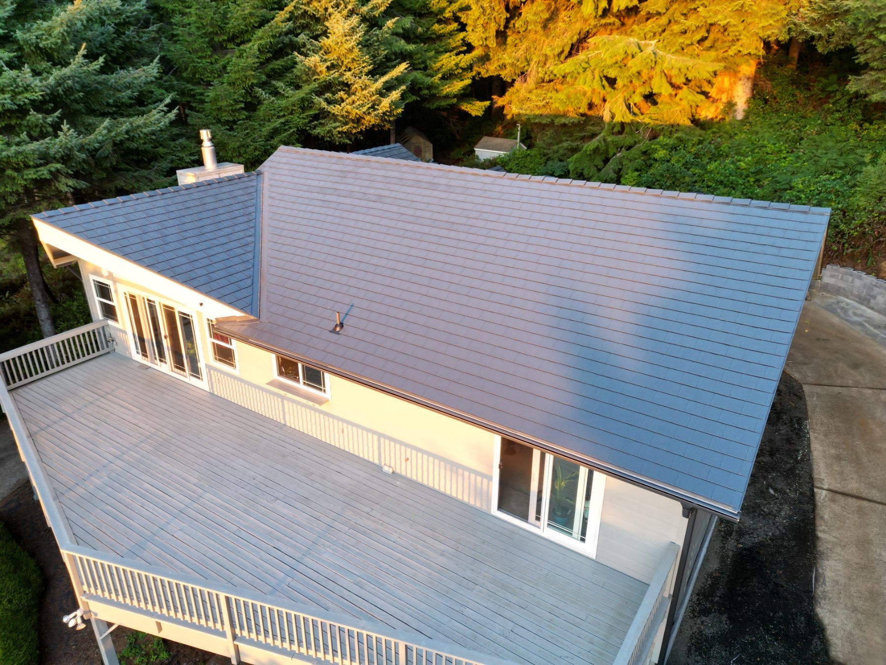 Aerial rear view of a 2-story ranch home with Interlock deep charcoal aluminum slate metal roofing, large deck, chimney, and forested backdrop in Yachats, OR.