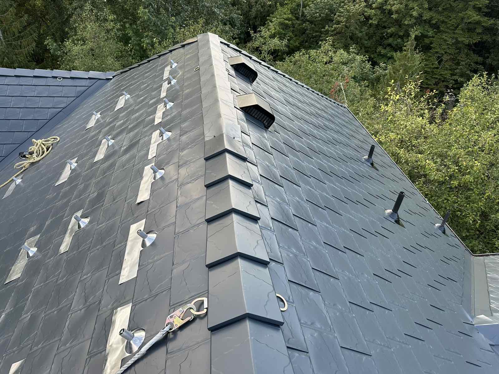Deep charcoal Interlock aluminum slate metal roofing with ridge cap detail, roof louvers, vent pipes, and safety anchors on a steep multi-hip roof surrounded by forest.