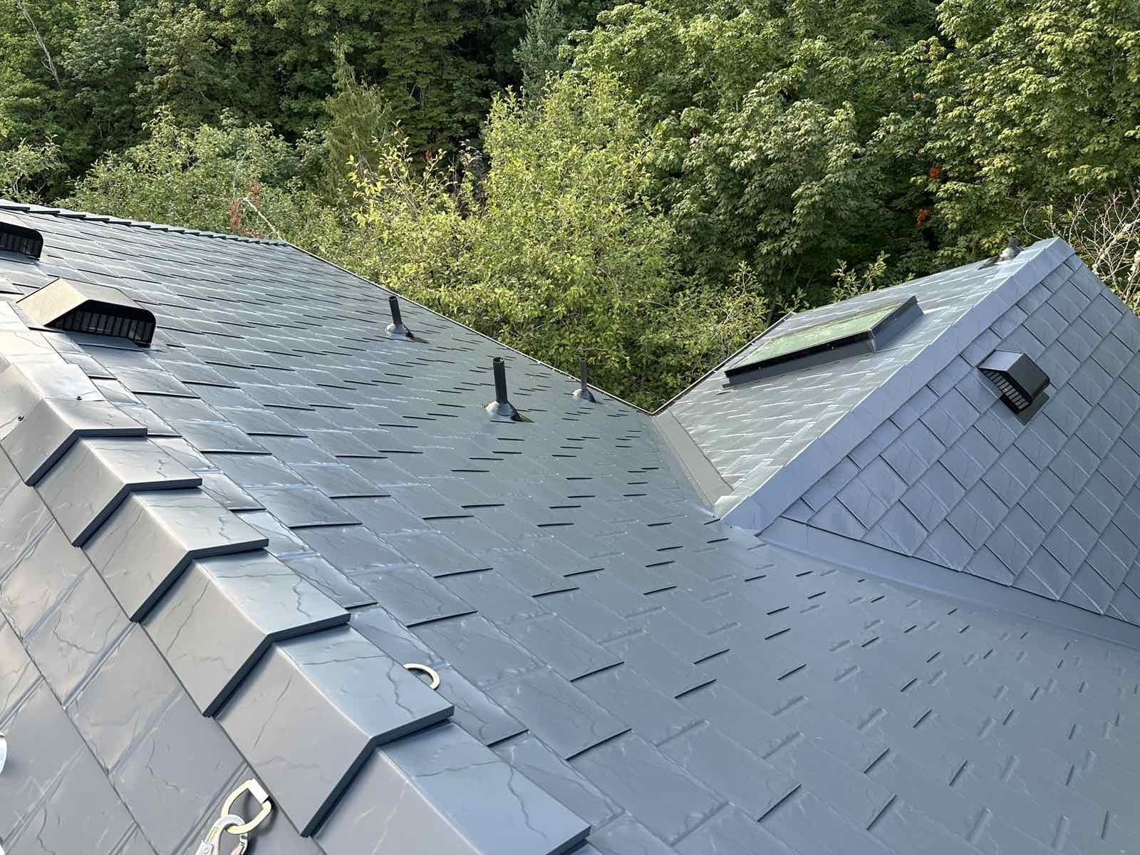 Aerial view of a complex multi-hip Deep Charcoal Interlock aluminum slate roof with valleys, vent pipes, and skylights surrounded by lush green trees.