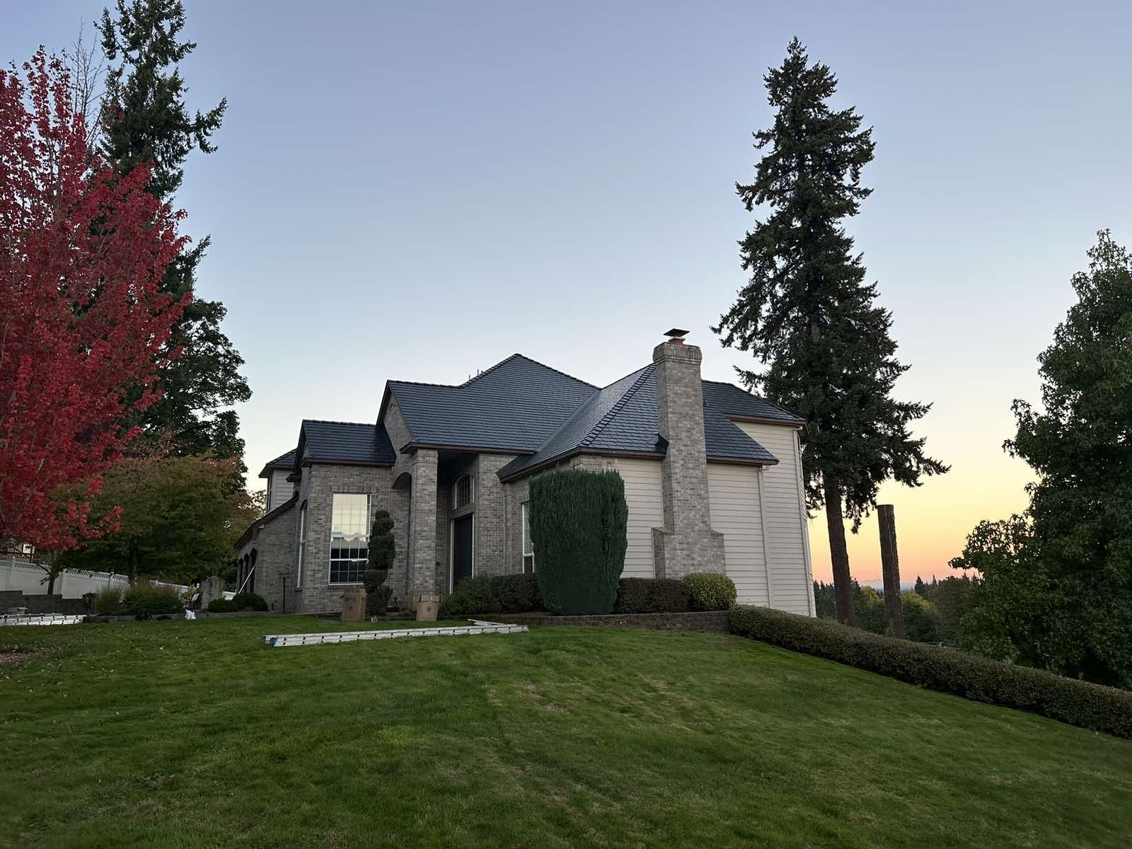 Large Tudor-style home with steep multi-hip black aluminum slate metal roof and stone chimney, set on a hillside at twilight in Scappoose, OR.