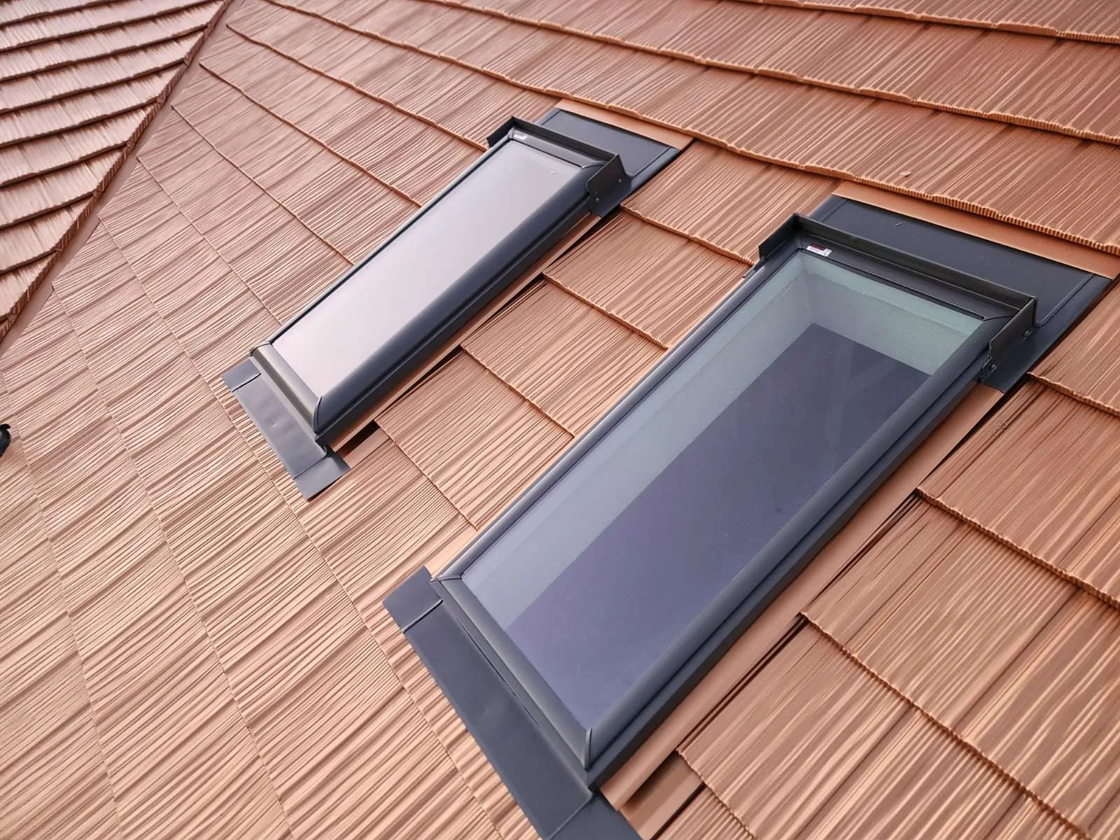 Close-up of two dark-framed skylights flush-mounted in Copper Penny Interlock aluminum shake metal roofing, with precise flashing detail.