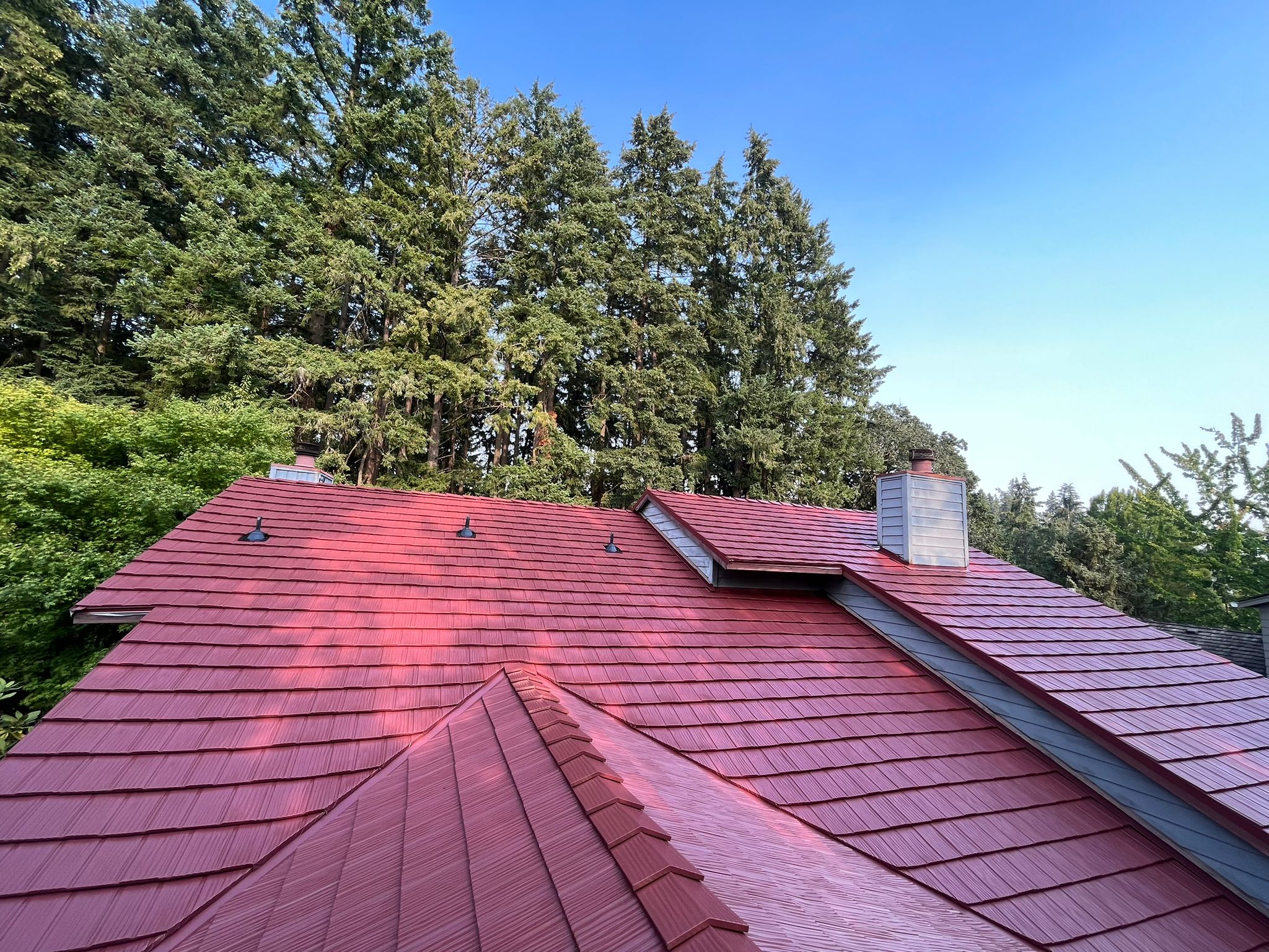 Tile red Interlock aluminum shake metal roof on a two-story Craftsman home in Beaverton OR, featuring complex multi-hip planes, valleys, dormers, and chimney amid tall evergreens.