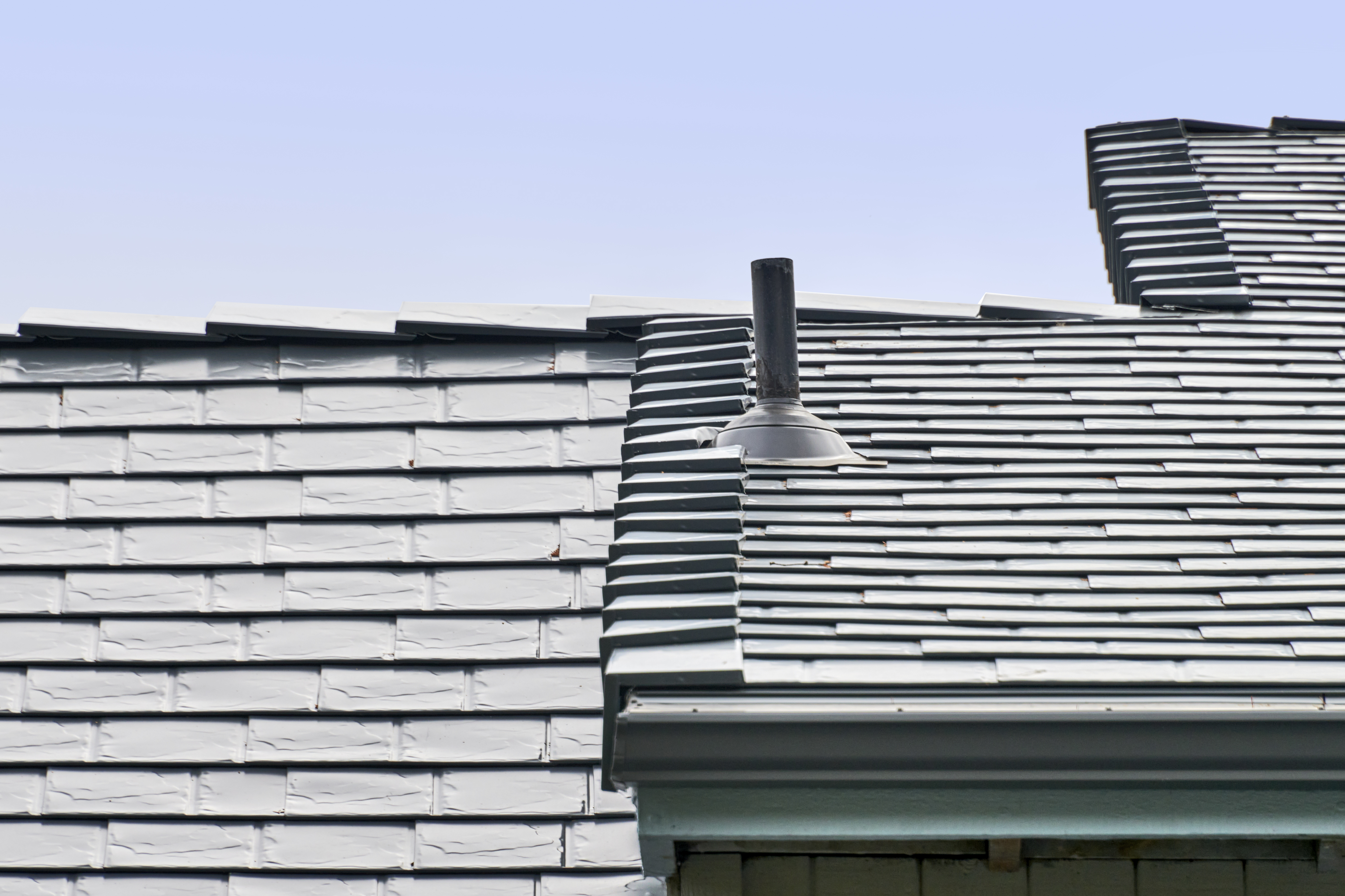 Close-up of Interlock deep charcoal aluminum slate roofing showing multi-level roof transition with vent pipe flashing detail against a clear blue sky.