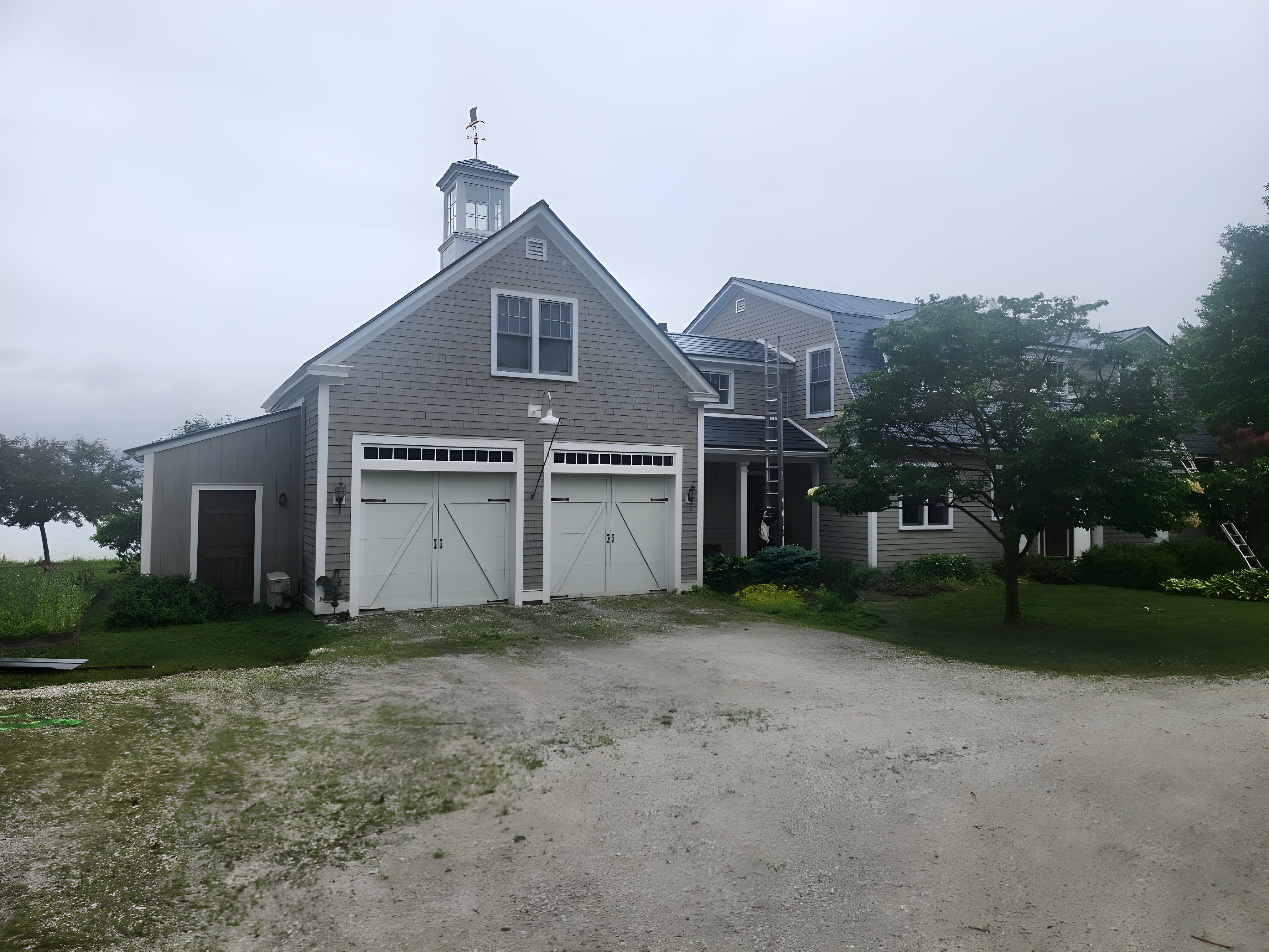 Two-story farmhouse with deep charcoal Interlock aluminum slate roof, cupola with weather vane, double white carriage garage doors, gray shingle siding, rural Vermont setting.