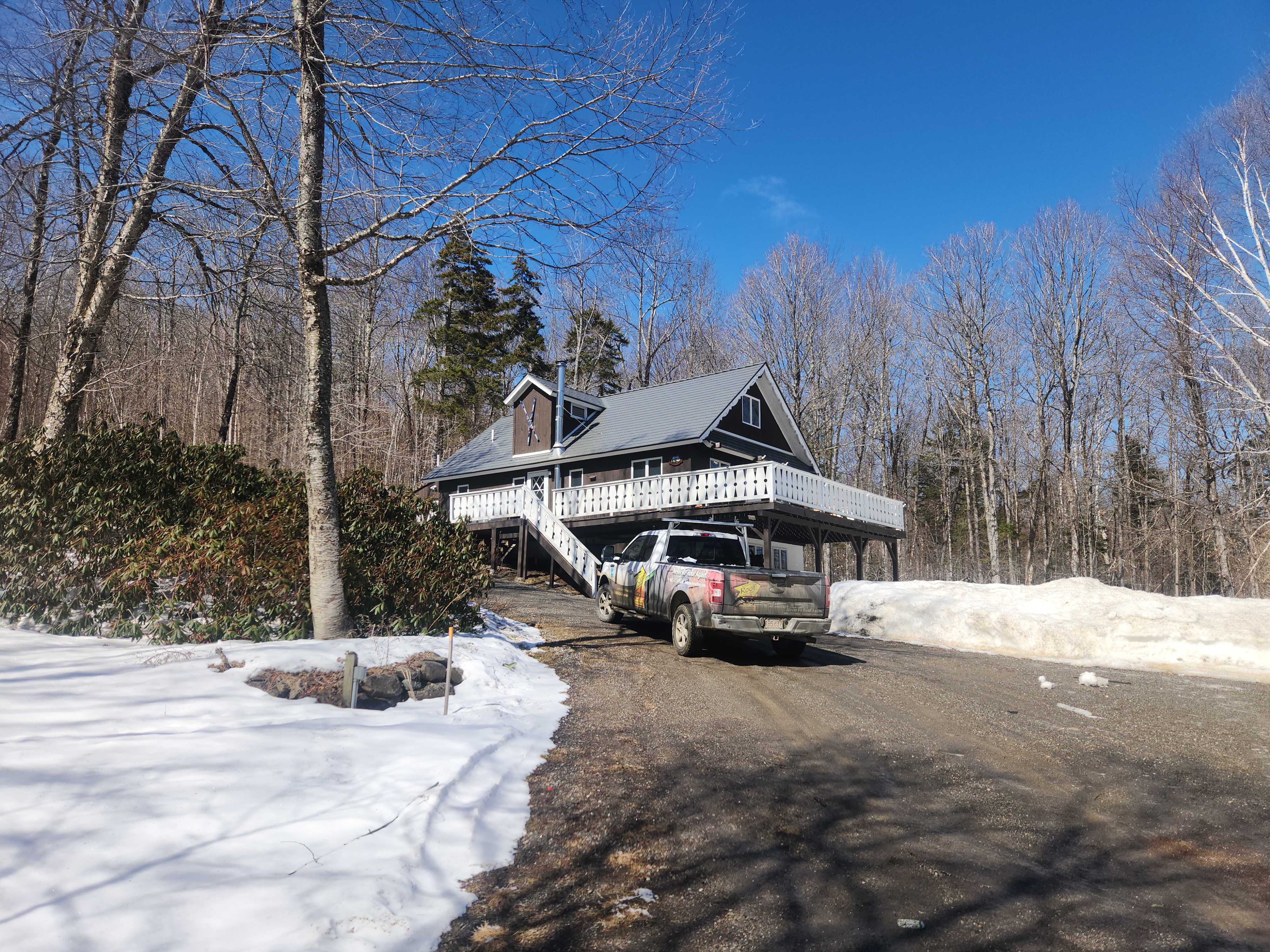 Chalet-style Vermont home with Interlock Musket Grey cedar shingle metal roof, steep gable profile, and white wraparound deck, surrounded by bare winter trees.