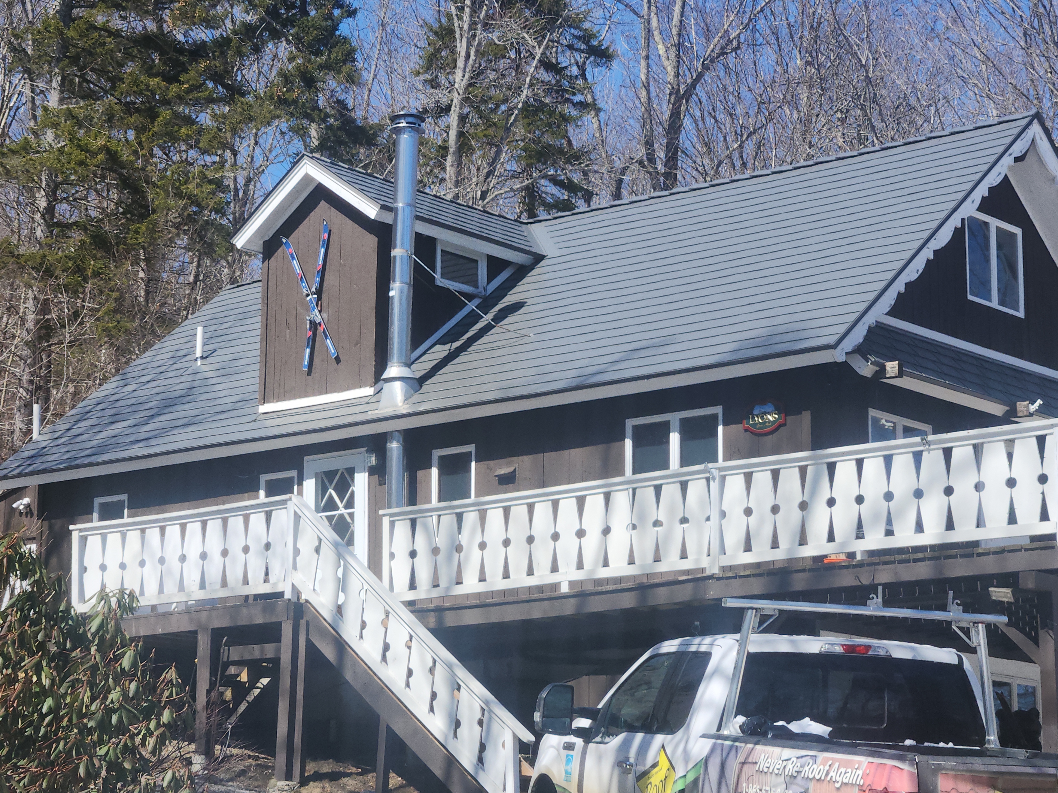 Two-story chalet-style Vermont home with Interlock Musket Grey cedar shingle aluminum roof, front dormer, vent pipe, and white deck railings amid winter forest.