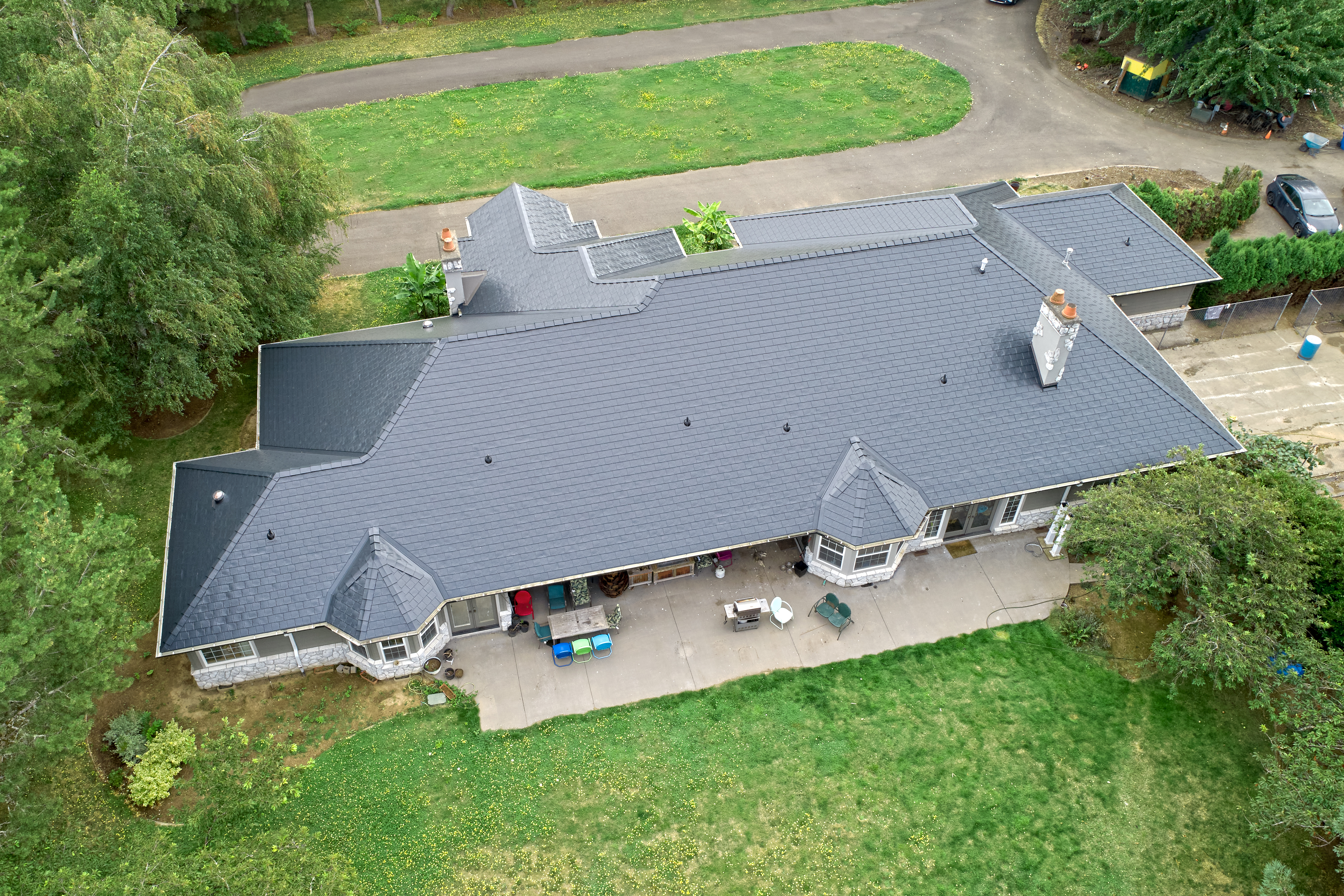 Aerial rear view of a large ranch-style home in Ridgefield, WA with a complex Interlock aluminum slate roof in charcoal black, featuring turreted dormers and two chimneys.