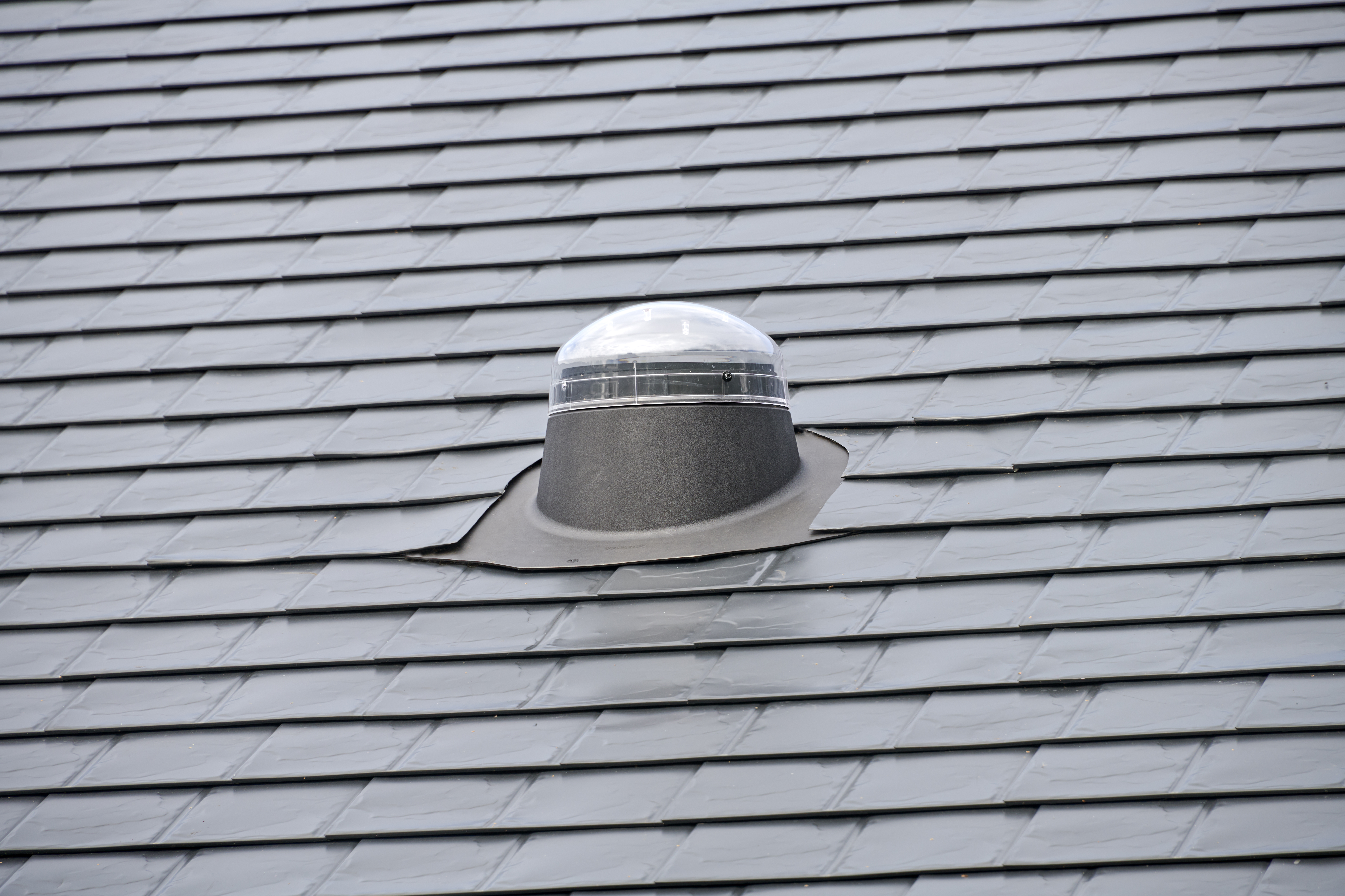 Close-up of a solar tube dome skylight with lead flashing installed on a dark grey Interlock aluminum slate metal roof.
