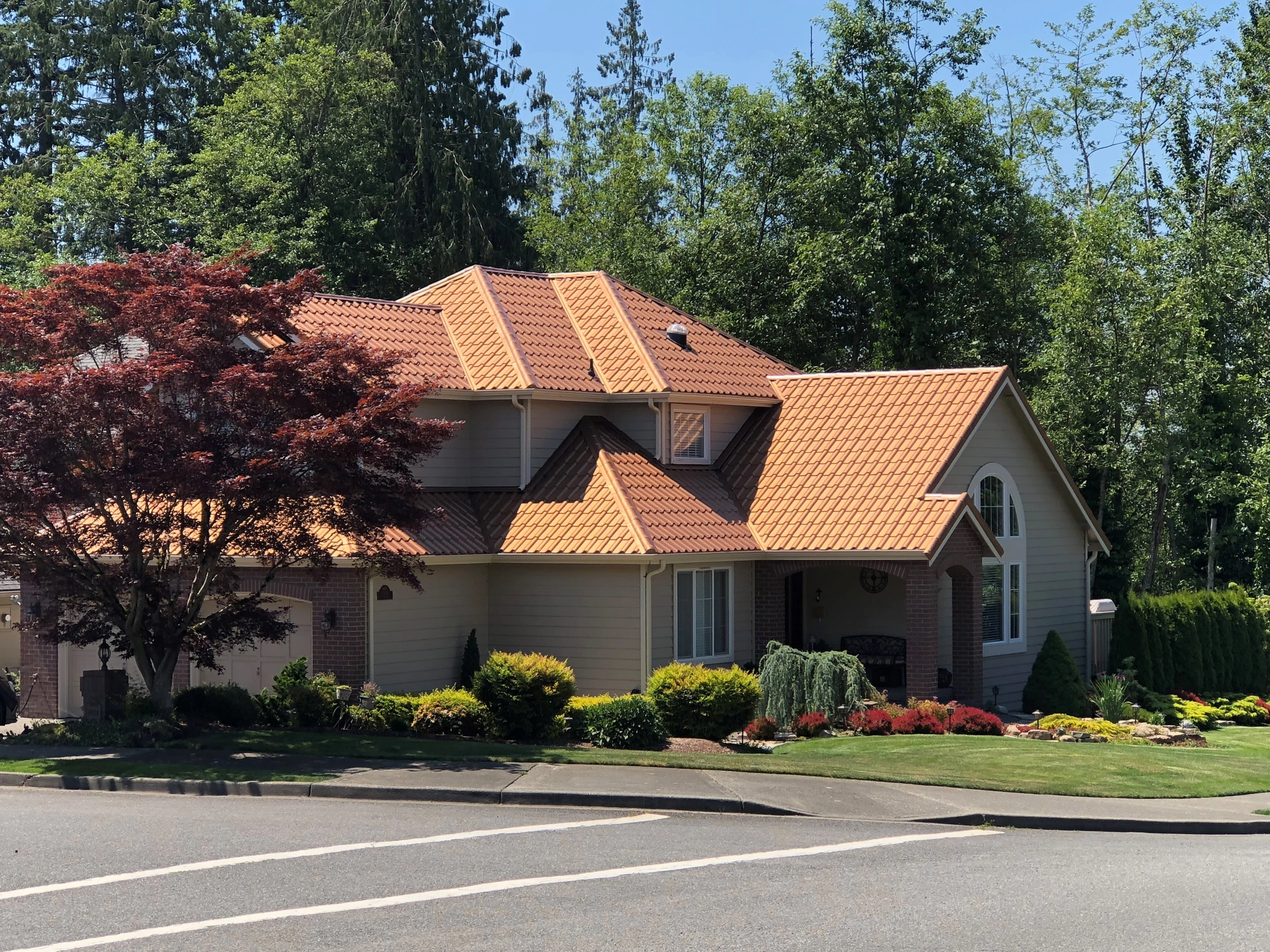 Aged Copper Interlock Tile aluminum roof on a tan two-storey home with dormers among tall trees, Arlington, Washington