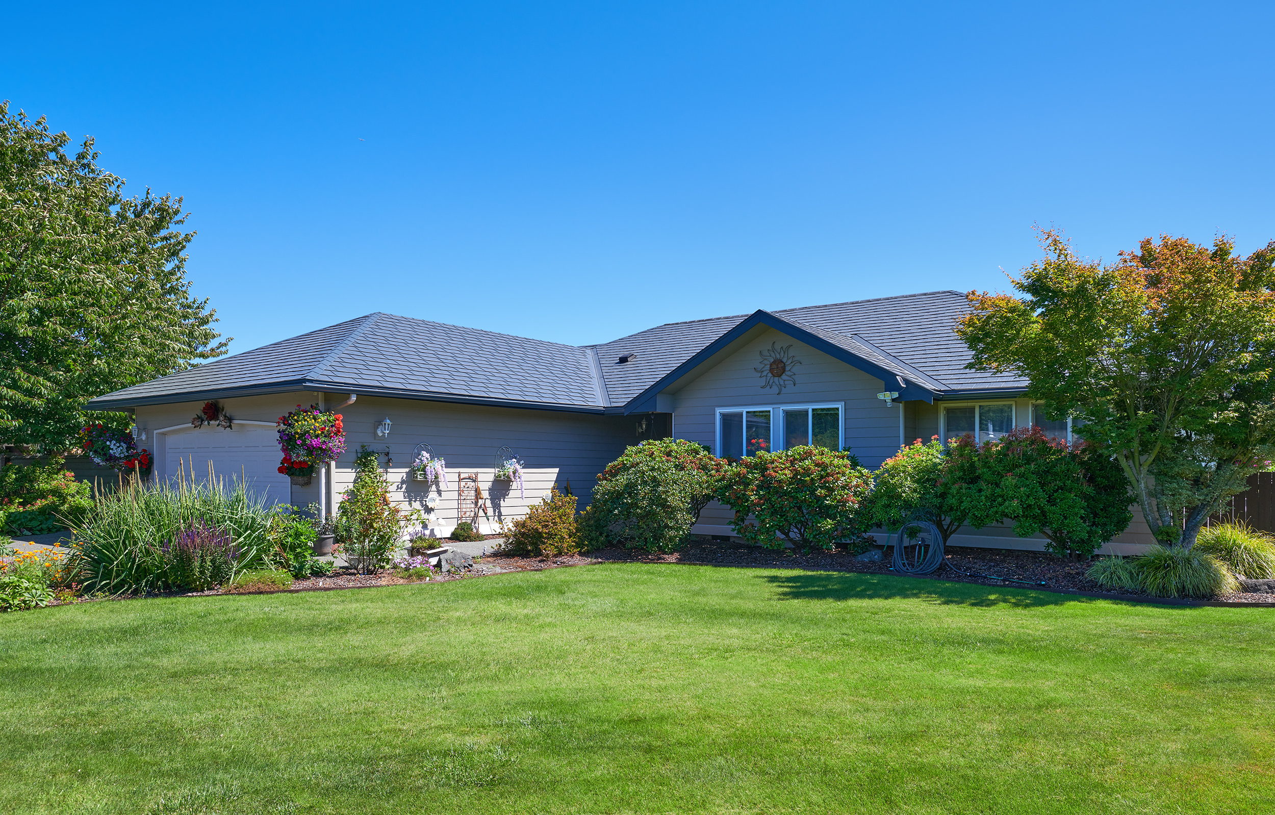 Single-story ranch home with Interlock deep charcoal aluminum slate roof featuring multi-hip complexity, lush manicured landscaping, and attached garage in Sequim, WA.